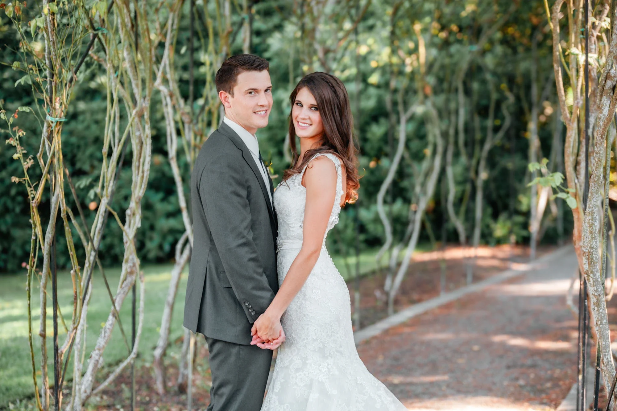A newlywed couple holding hands and smiling at the camera outdoors, surrounded by trees and a garden pathway.