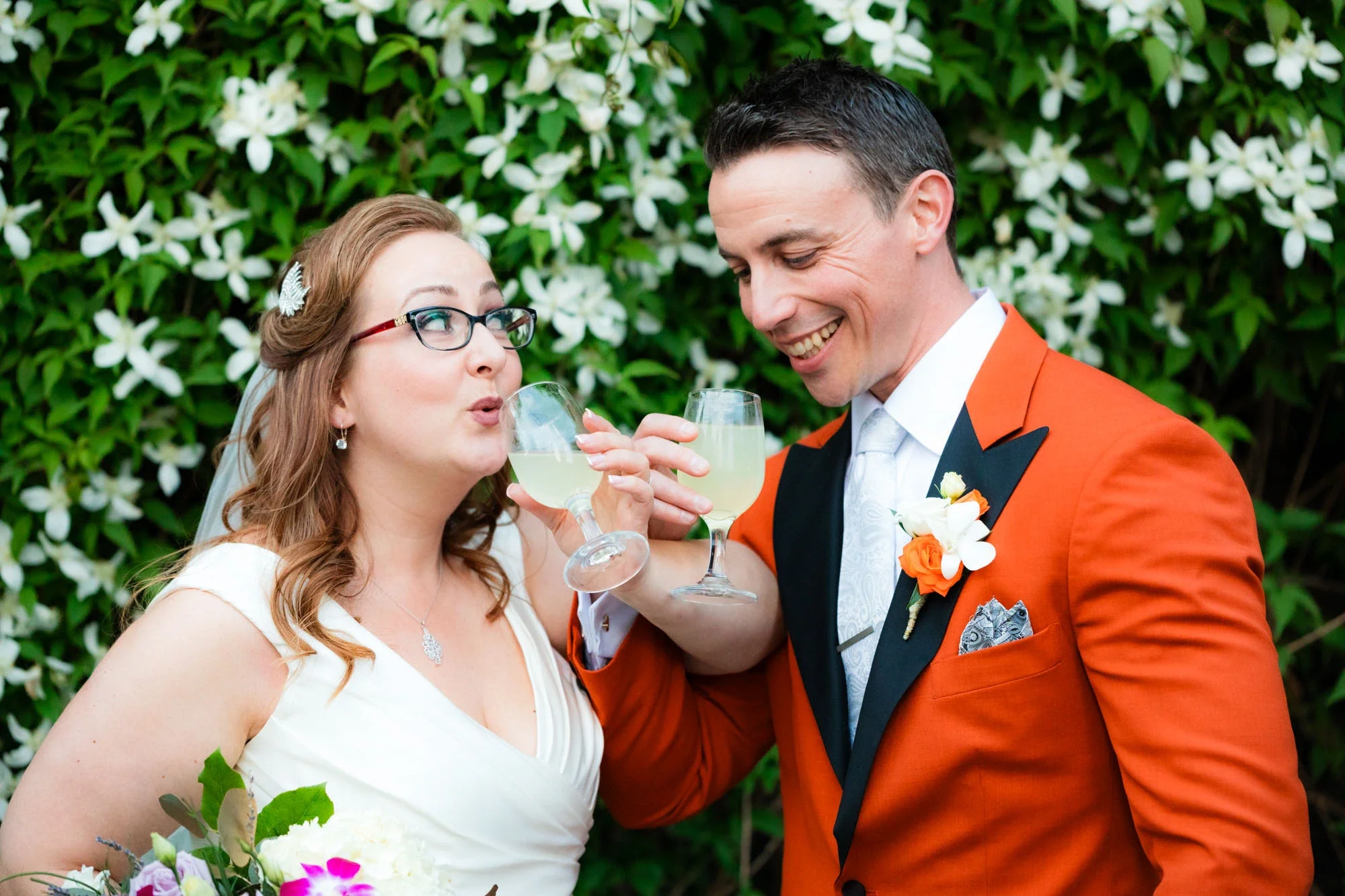 Bride and groom sharing a playful champagne toast surrounded by lush white blossoms, groom in bold orange suit with matching boutonniere and bride in elegant white gown with veil and colorful bouquet, affordable wedding photography by Shakil Hussain 