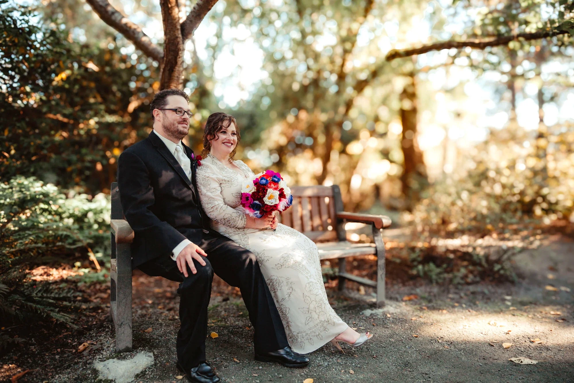 Romantic bride and groom sharing a joyful moment while seated on a wooden bench in a sunlit Abbotsford forest. The bride holds a vibrant multicolored bouquet, with dreamy bokeh lights filtering through the trees. Captured by Shakil Hussain, Abbotsfor
