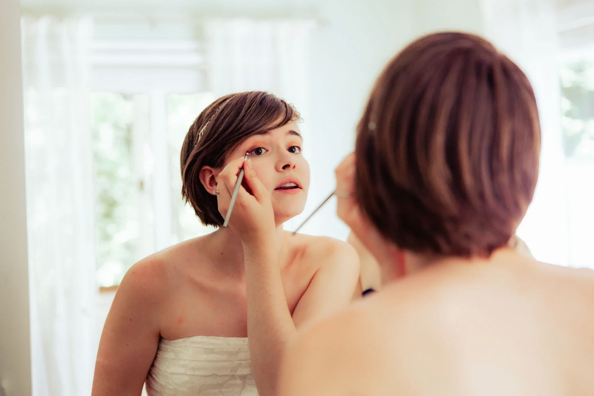 Bride Danika having her makeup applied while looking in the mirror during bridal preparations, wearing a strapless wedding gown with delicate hair accessory and soft natural window light illuminating the getting-ready moment, affordable wedding photo