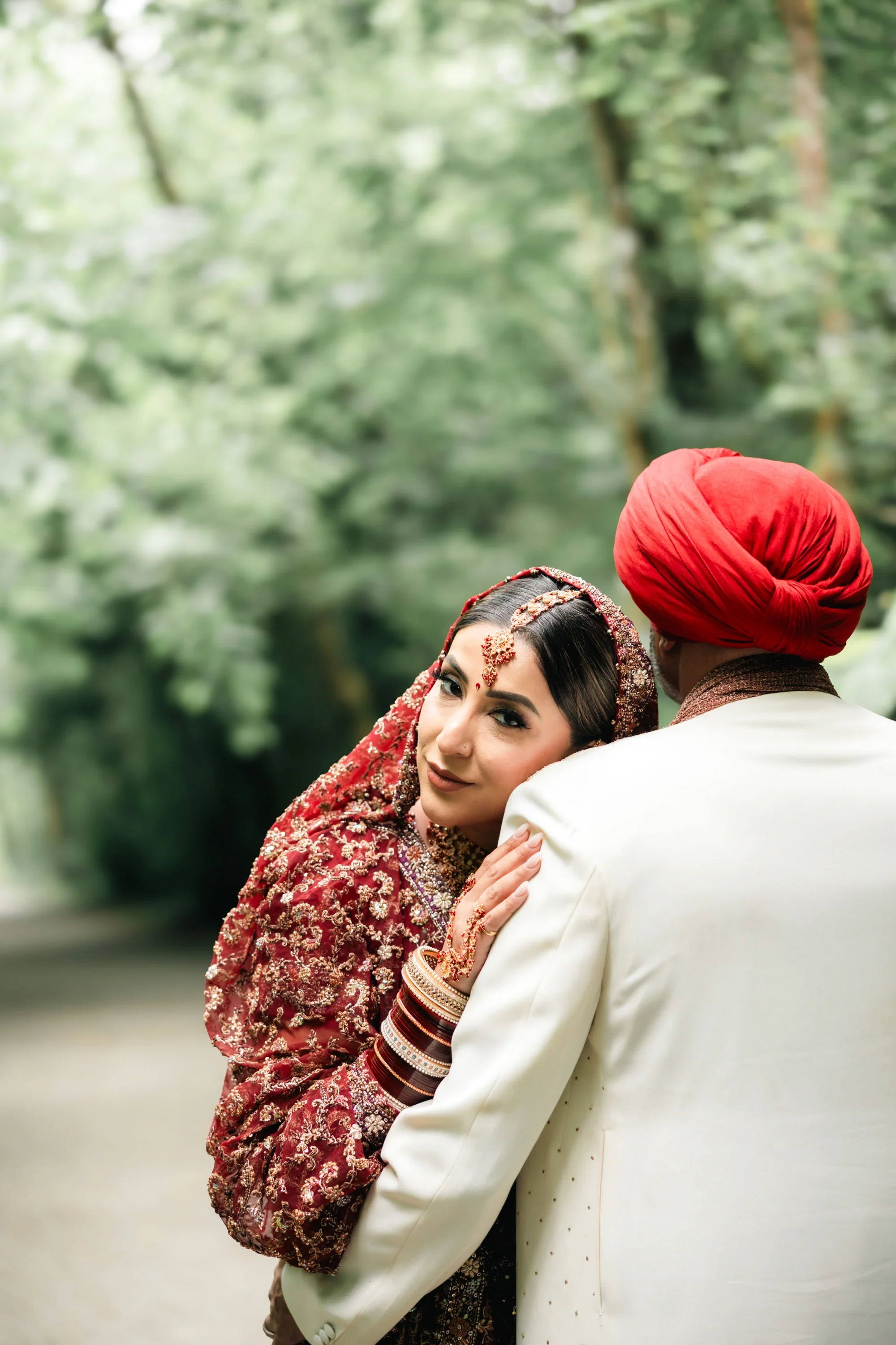 Tender embrace between bride and groom in traditional Indian wedding attire on a serene forest path, with bride in richly embroidered maroon lehenga and groom in cream sherwani with red turban looking lovingly at her, affordable wedding photography b