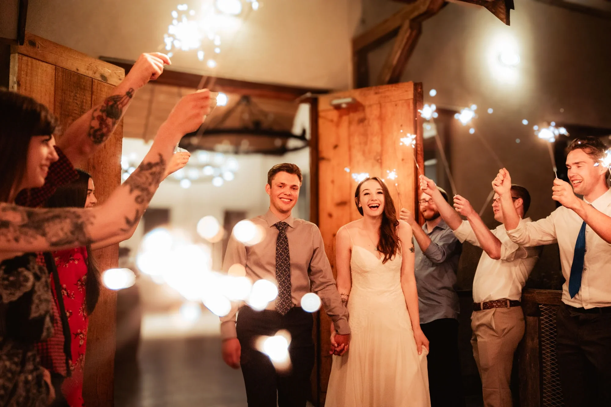 Joyful bride and groom making a magical sparkler exit at their wedding reception, surrounded by smiling guests holding sparklers under warm bokeh lights in a rustic venue, affordable wedding photography by Shakil Hussain Photography serving Abbotsfor