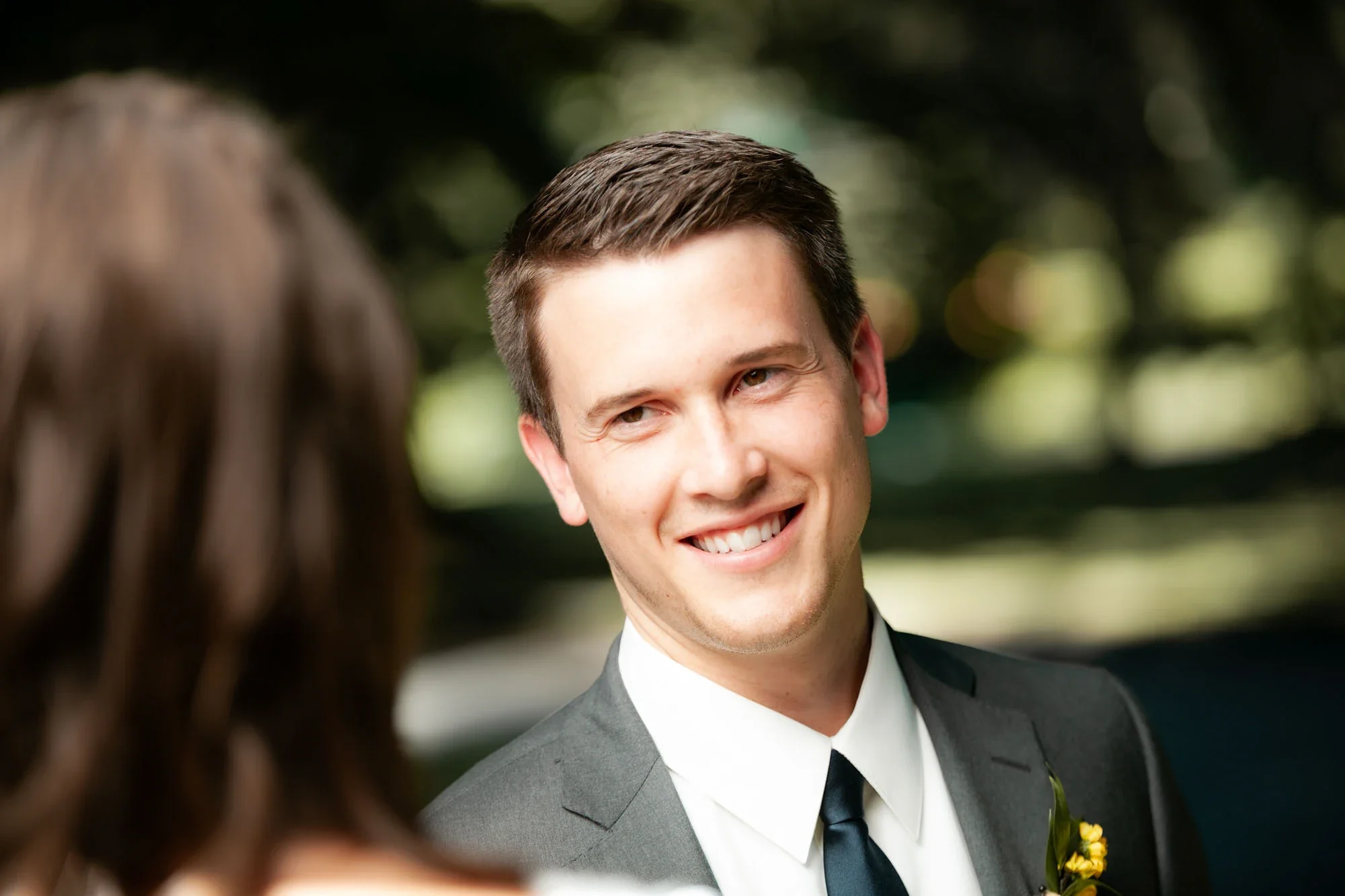 Handsome groom in charcoal gray suit with white shirt, navy tie, and yellow boutonniere smiling warmly during the wedding ceremony, soft natural light highlighting his joyful expression, affordable wedding photography by Shakil Hussain Photography se
