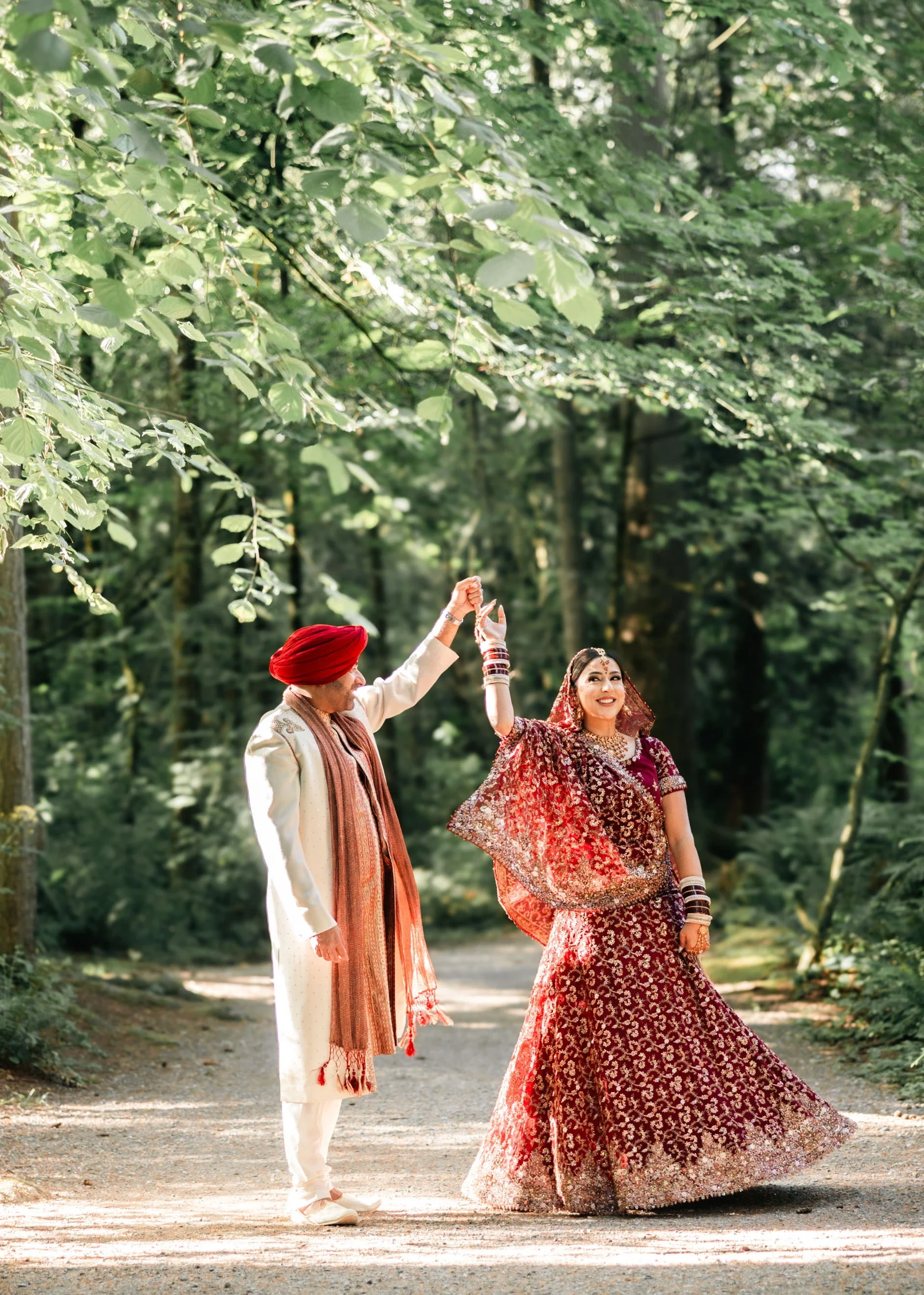 Vibrant Indian bride and groom in traditional attire joyfully dancing hand-in-hand on a forest path surrounded by lush green trees, with red turban and embroidered maroon lehenga, captured by an Abbotsford Fraser Valley Vancouver wedding photographer