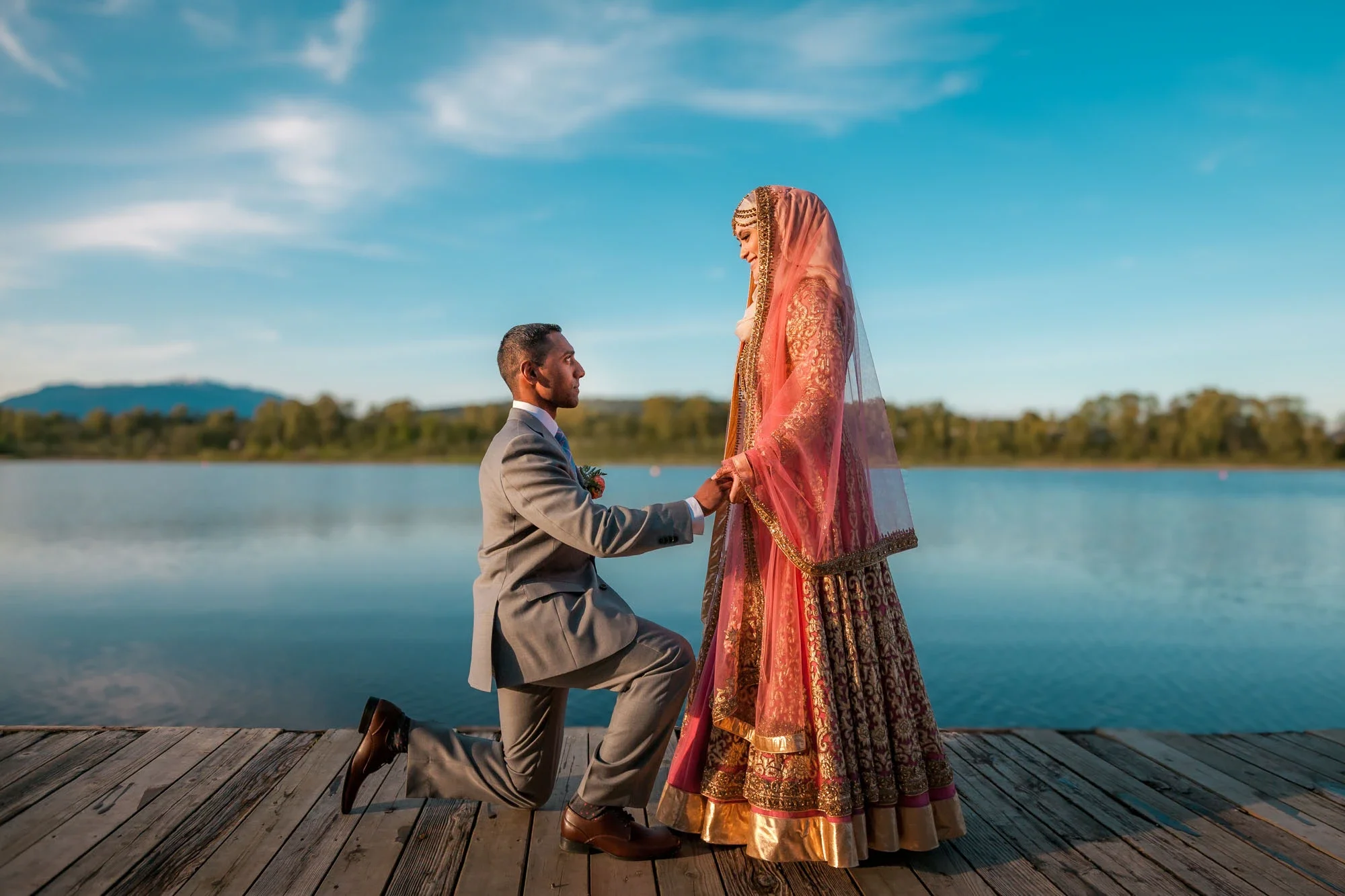 Romantic fusion wedding portrait with groom in gray suit kneeling on one knee holding hands with bride in traditional pink and gold embroidered Indian lehenga on a wooden dock overlooking a serene lake under blue skies, affordable wedding photography