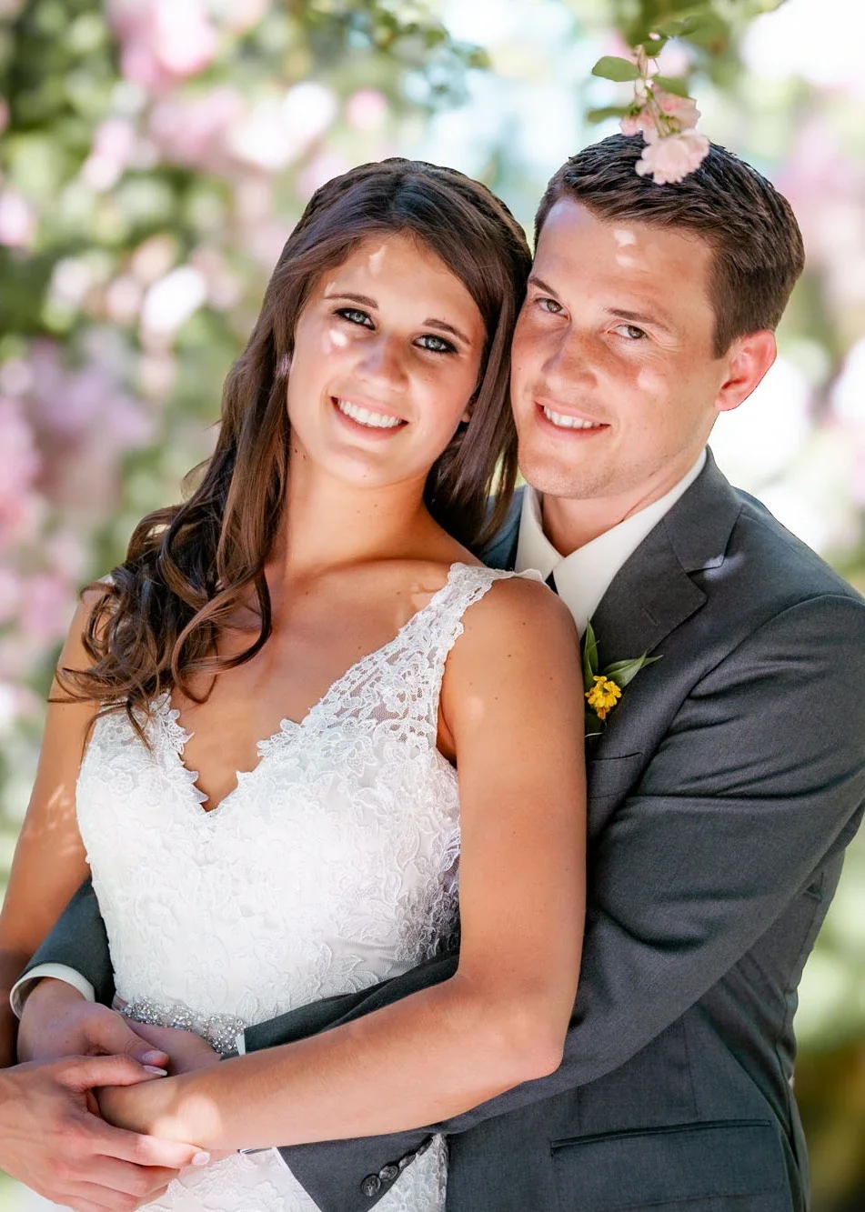 Joyful bride Elizabeth and her groom embracing and smiling in front of soft pink spring blossoms, with bride in a lace V-neck wedding gown and groom in charcoal suit with yellow boutonniere, photographed by a professional Abbotsford Fraser Valley Van
