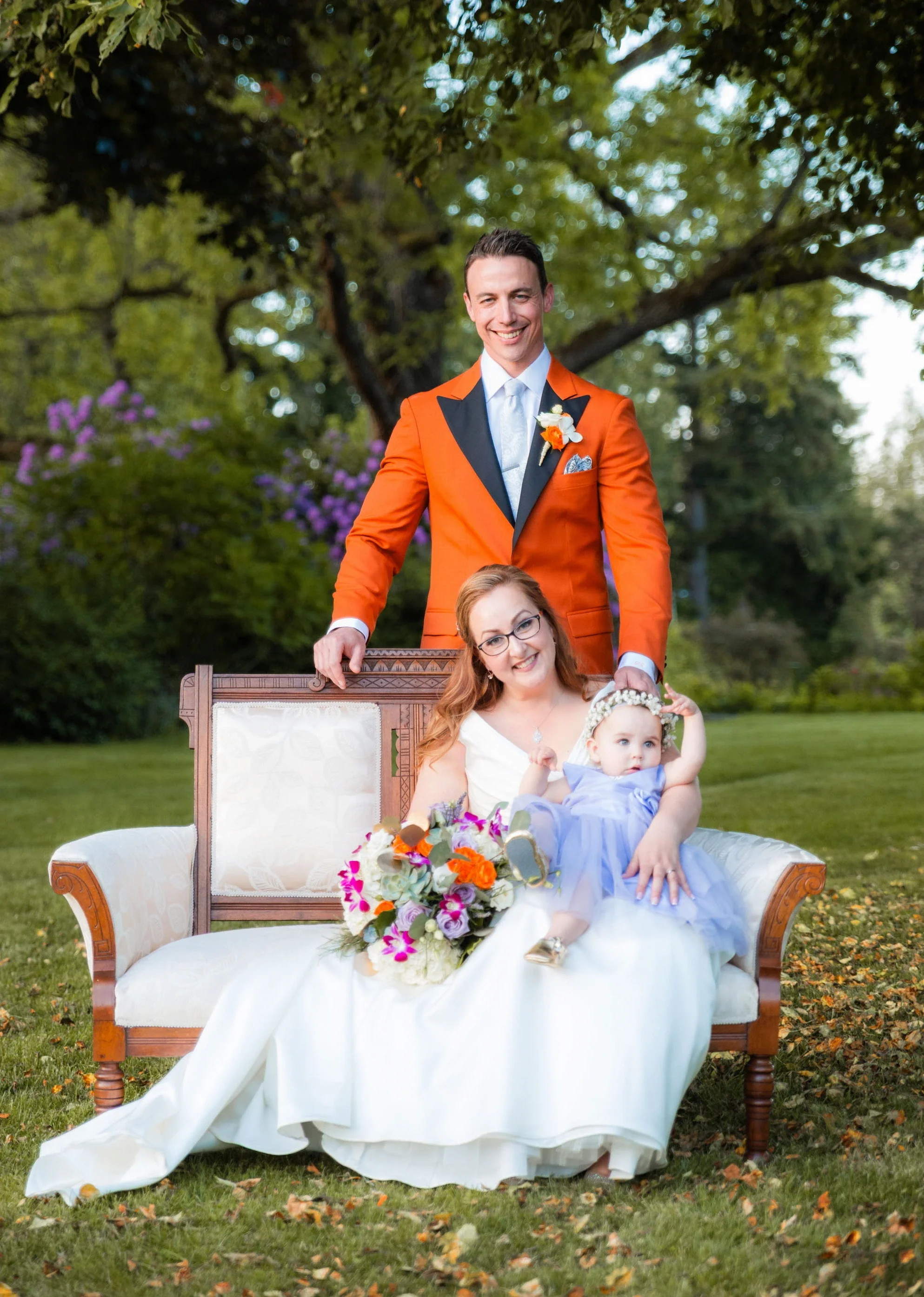 Joyful bride and groom with their baby daughter in a sweet family portrait, seated on a vintage bench in a lush garden setting with vibrant orange suit and colorful bouquet, captured by an Abbotsford Fraser Valley Vancouver wedding photographer.