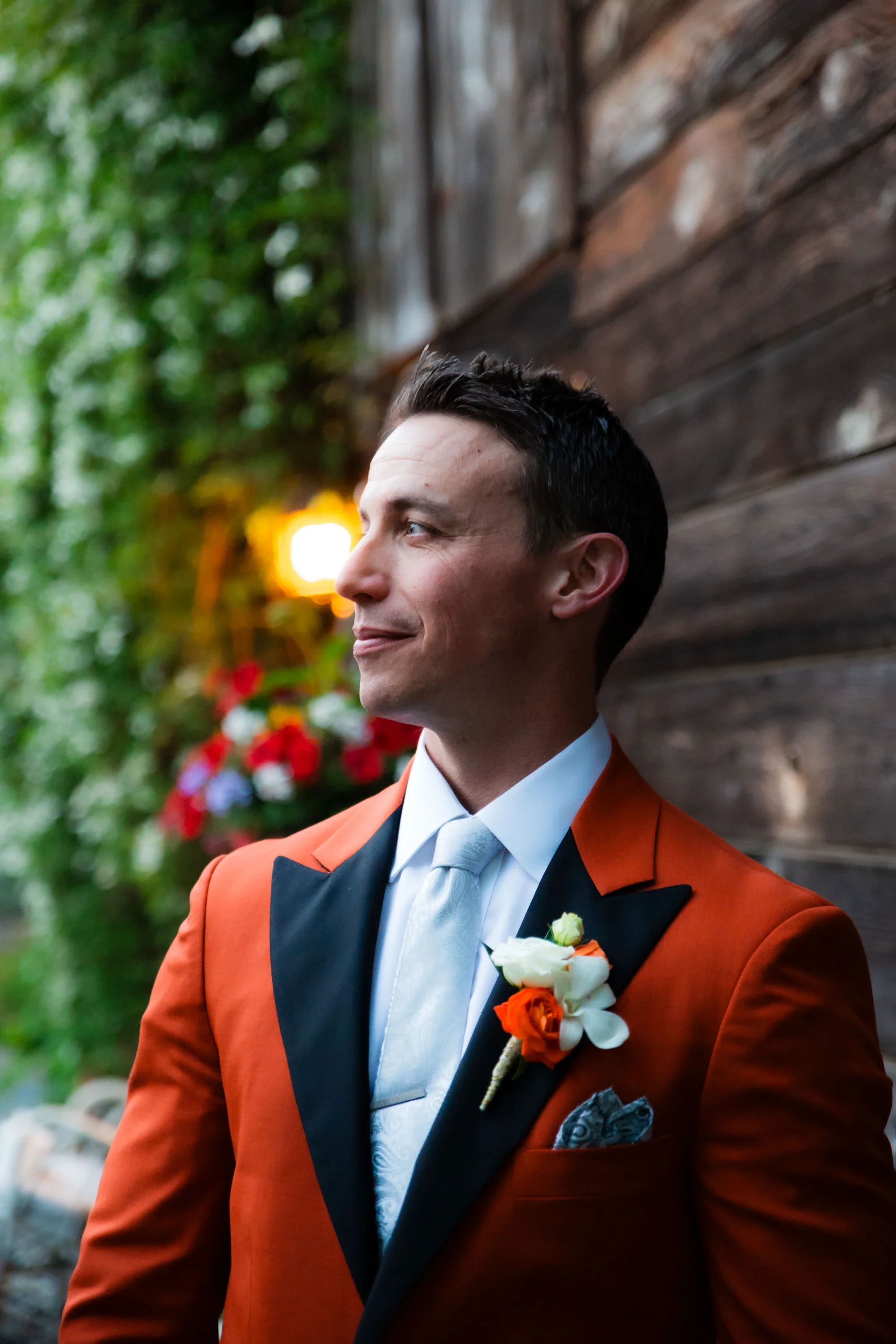Stylish groom in bold orange suit jacket with black lapels, silver tie, and vibrant orange and white rose boutonniere looking thoughtfully to the side against a rustic wooden wall and blurred greenery, affordable wedding photography by Shakil Hussain