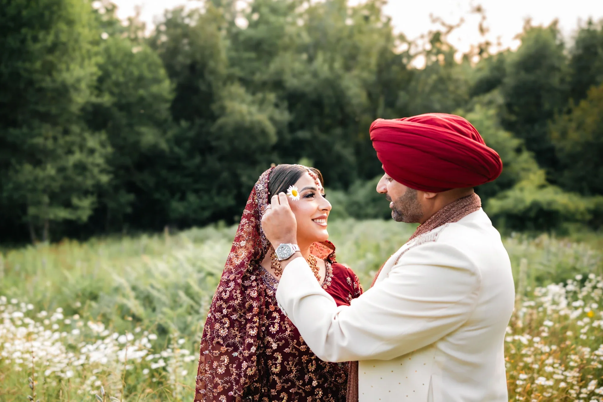A couple dressed in traditional Indian wedding attire sharing a joyful moment outdoors among greenery and flowers.