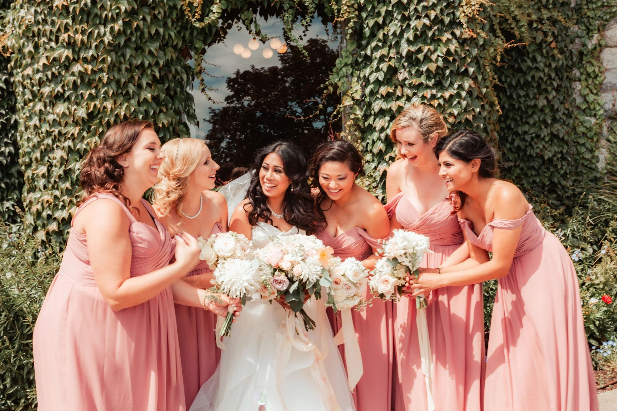 Joyful bride surrounded by her smiling bridesmaids in matching blush pink dresses, all holding elegant white and pastel bouquets, gathered under a lush ivy-covered arch in a romantic garden setting, affordable wedding photography by Shakil Hussain Ph