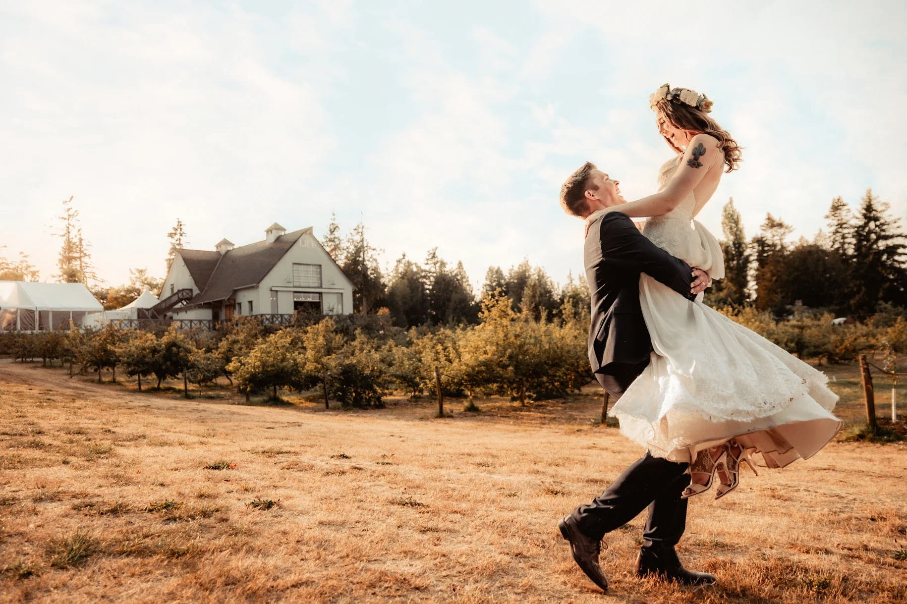 A groom lifting a bride in a field with a white house and trees in the background during sunset.