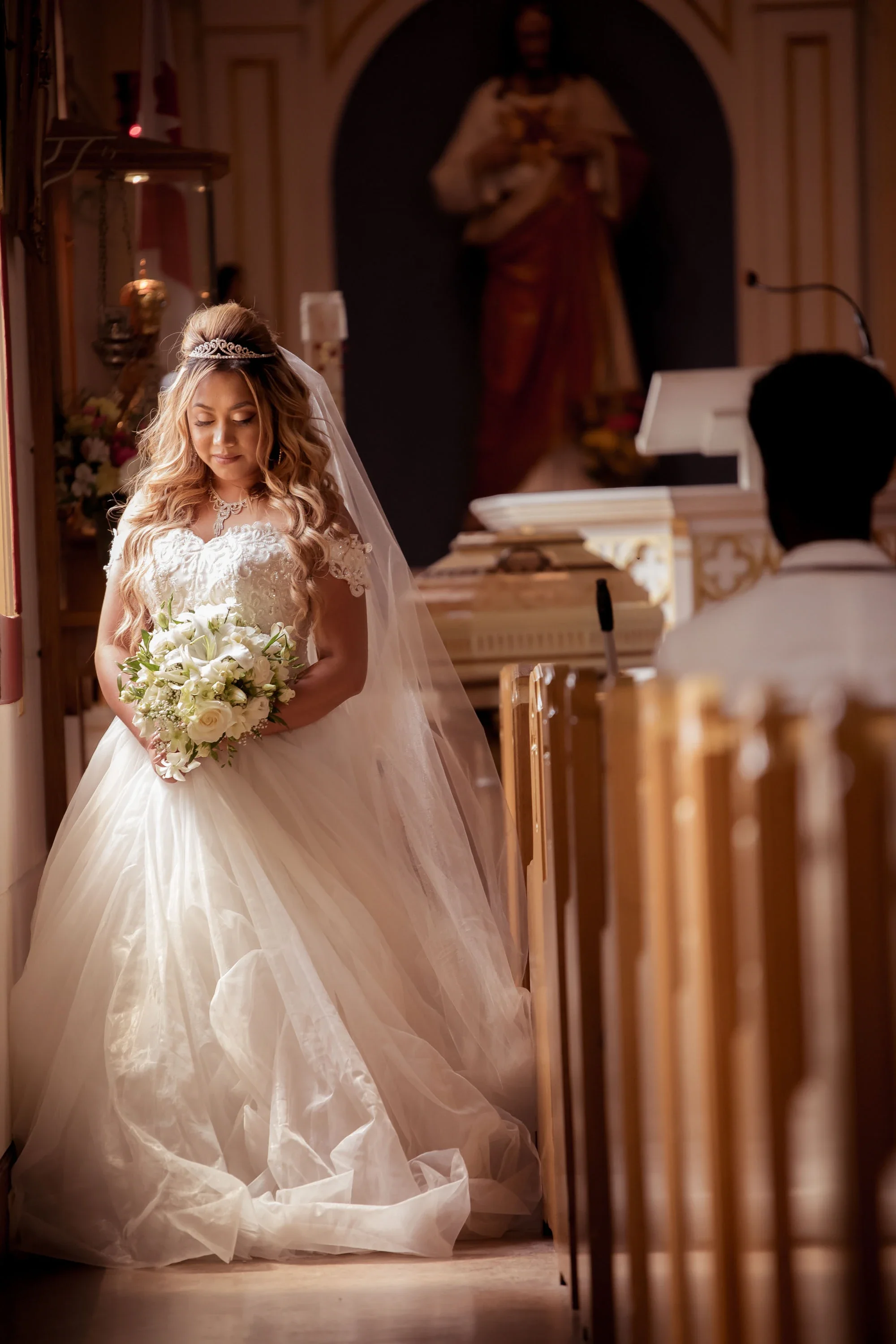 Serene bride in a long-sleeved lace wedding gown with flowing tulle skirt and veil, holding a elegant white lily and rose bouquet, standing thoughtfully in a softly lit Catholic church with a statue of Jesus in the background, affordable wedding phot