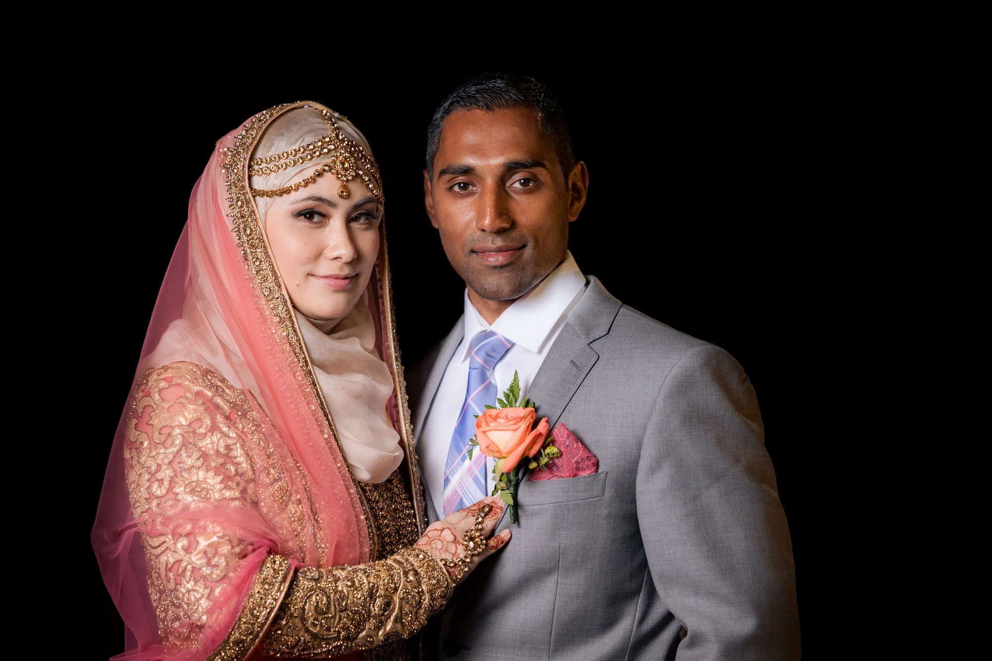 Elegant Muslim bride and groom in studio portrait against black background, bride in ornate pink and gold embroidered dress with hijab and henna hands touching groom's orange rose boutonniere, groom in gray suit with blue tie, affordable wedding phot