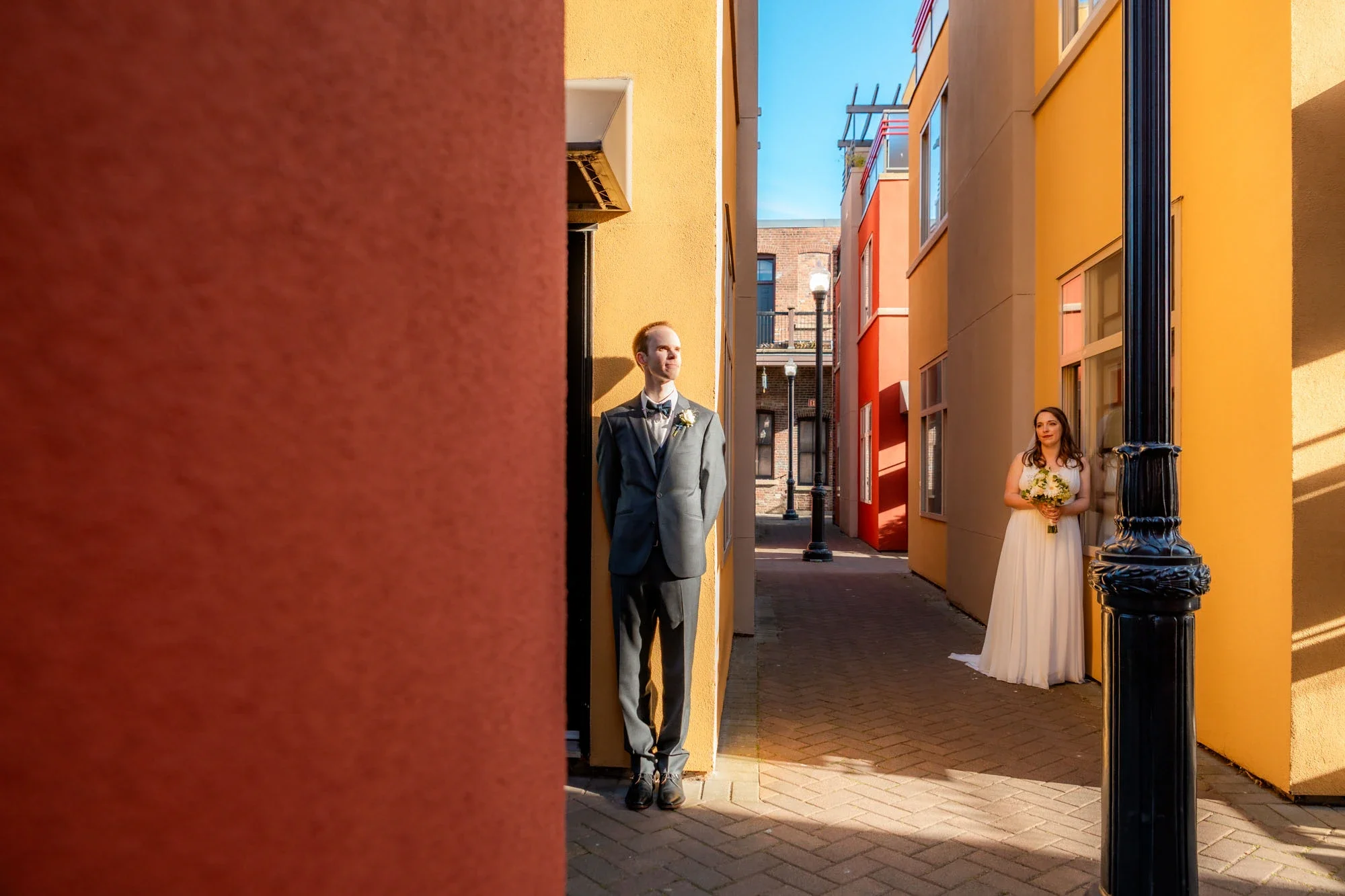Creative first look moment with groom in gray suit leaning against a red wall in a vibrant colorful alley, smiling toward the bride in a flowing white gown holding her bouquet at the opposite end in Abbotsford BC. Captured by Shakil Hussain, Abbotsfo