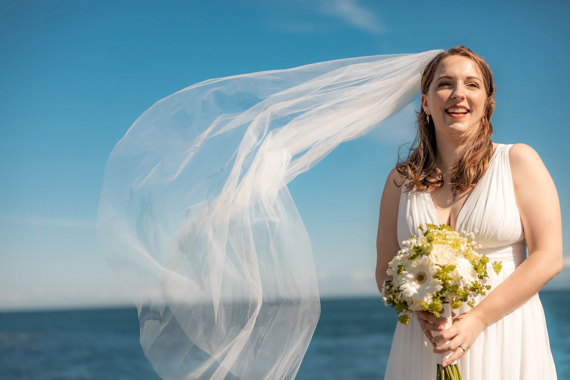 Radiant bride smiling joyfully as her long veil flows dramatically in the wind against a bright blue ocean and sky backdrop in Abbotsford BC, holding a white and green floral bouquet. Captured by Shakil Hussain, Abbotsford BC wedding photographer.