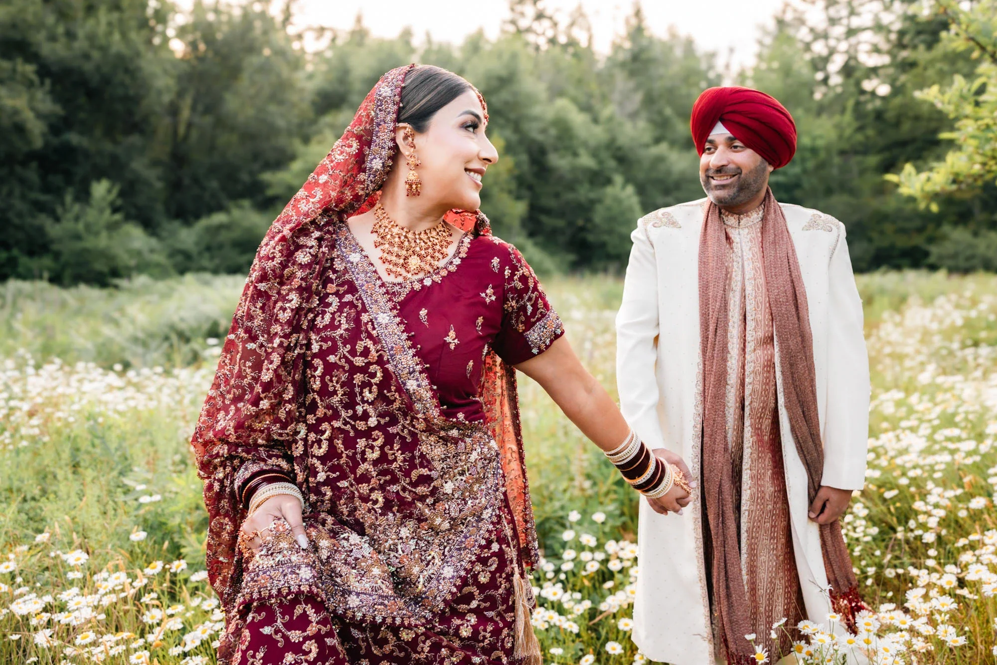 Bride and groom in traditional Indian attire holding hands and smiling while walking through a lush field of white wildflowers, with bride in richly embroidered maroon lehenga and groom in cream sherwani with red turban, affordable wedding photograph