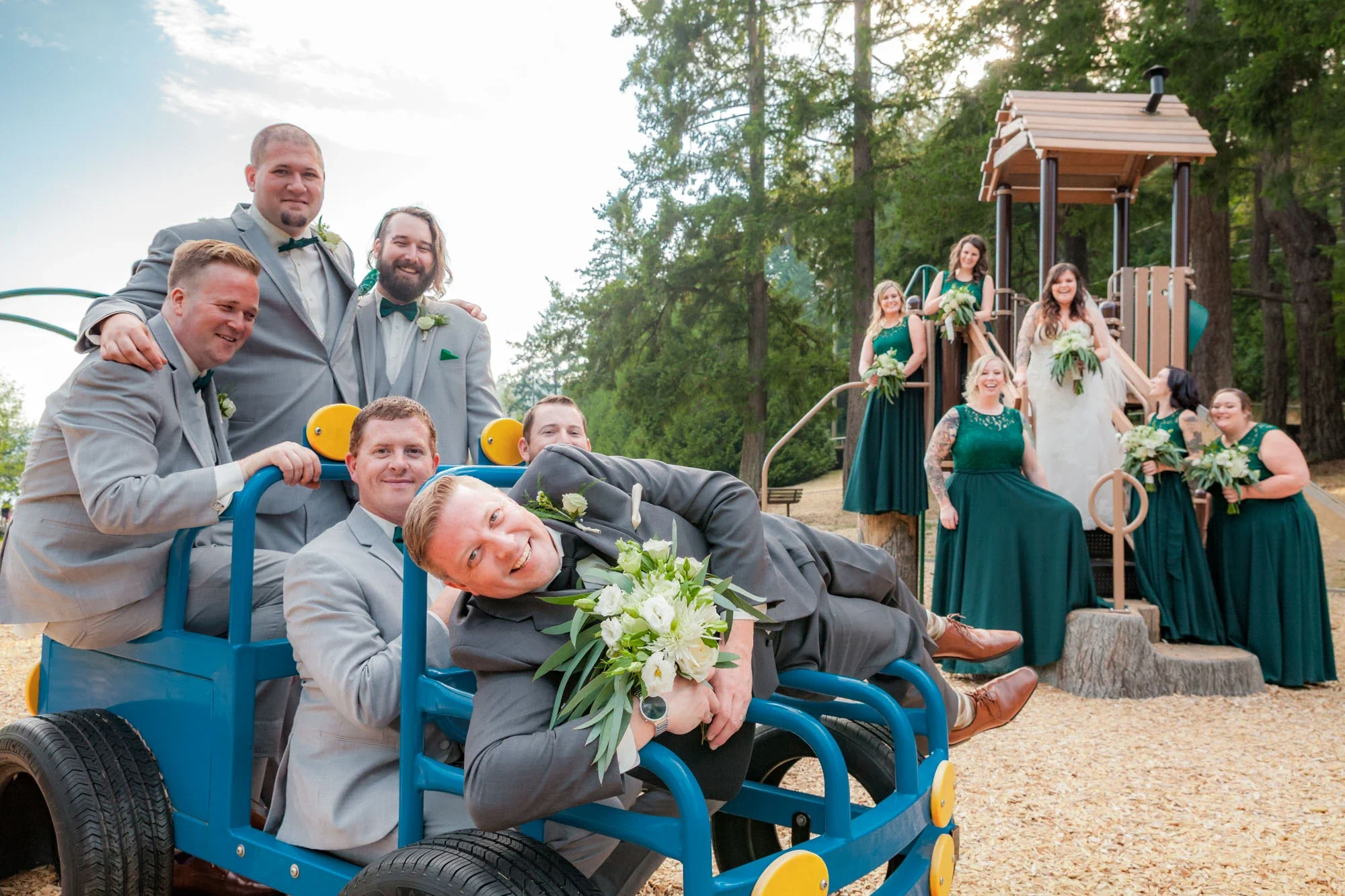 Fun and playful wedding party group portrait at a playground in Abbotsford BC, with groomsmen piled into a blue play structure holding the bride's bouquet and bridesmaids in emerald green dresses posing on the equipment. Captured by Shakil Hussain, A