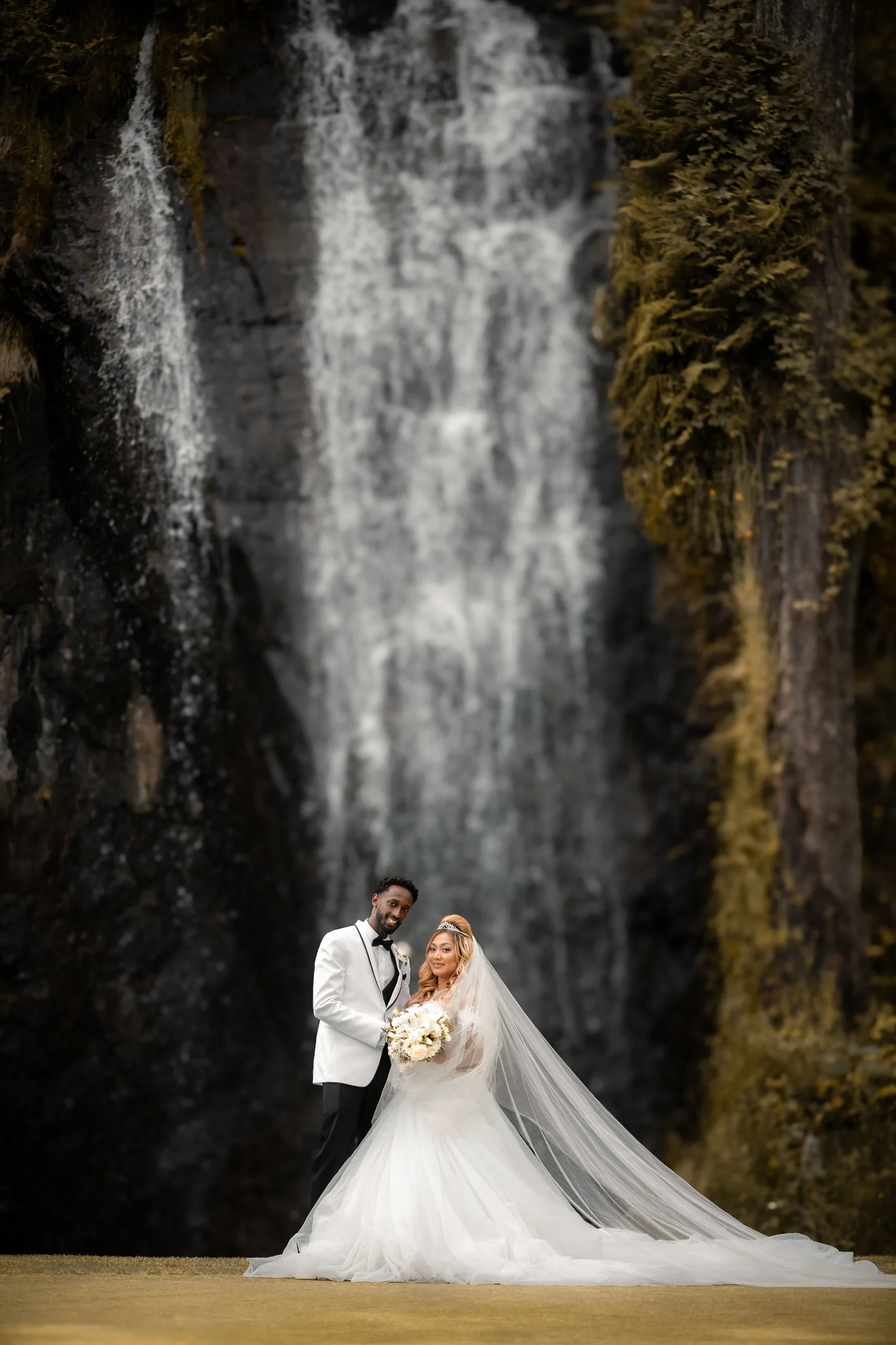 Romantic bride and groom portrait in front of a dramatic cascading waterfall surrounded by lush greenery, groom in white tuxedo jacket embracing bride in flowing ballgown with long veil and white bouquet, affordable wedding photography by Shakil Huss
