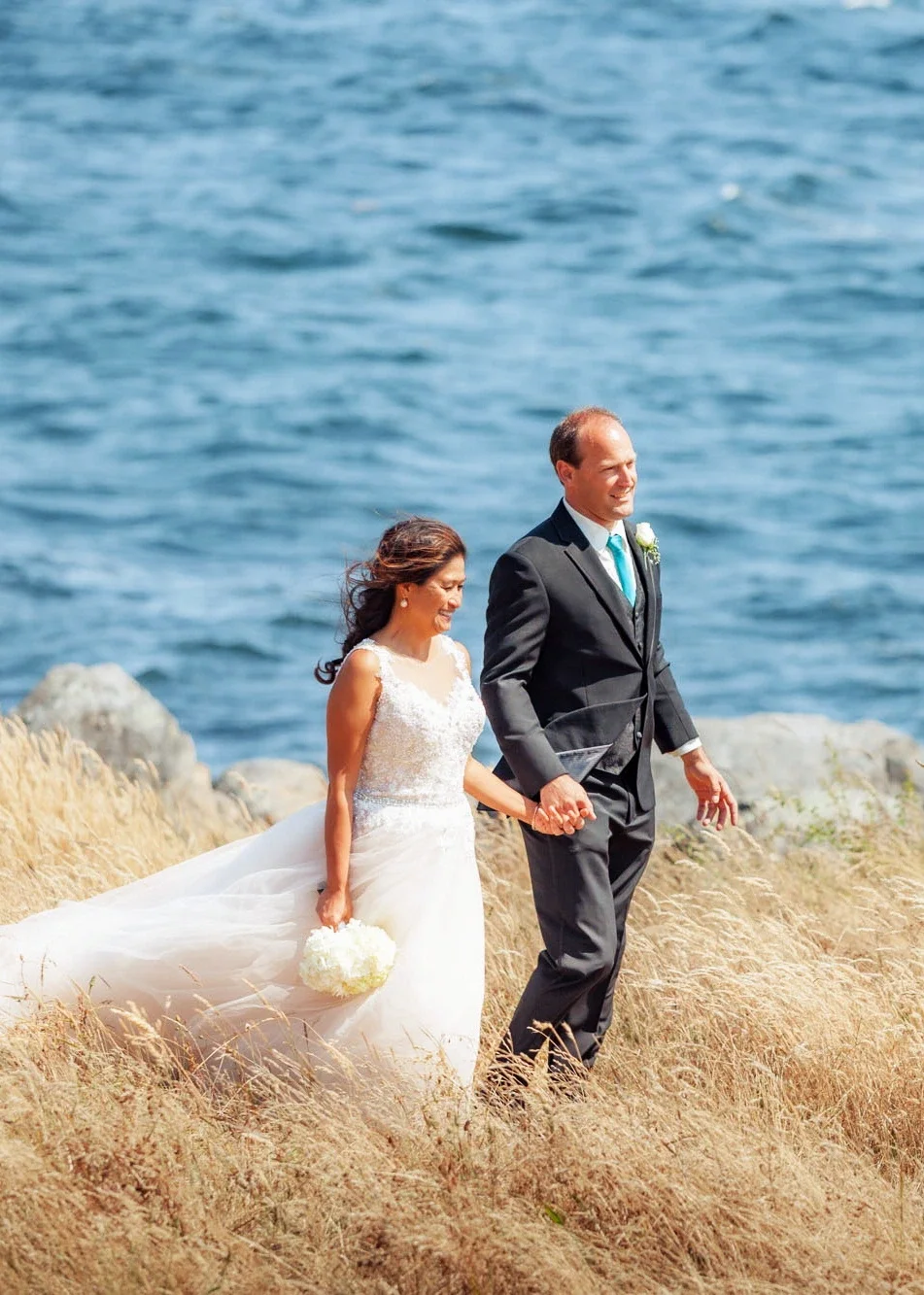 Happy bride Lotis and groom Bob holding hands and walking along a scenic ocean coastline with tall grasses and blue water, bride in a sparkling beaded wedding gown holding a white hydrangea bouquet and groom in classic black suit with teal tie, affor