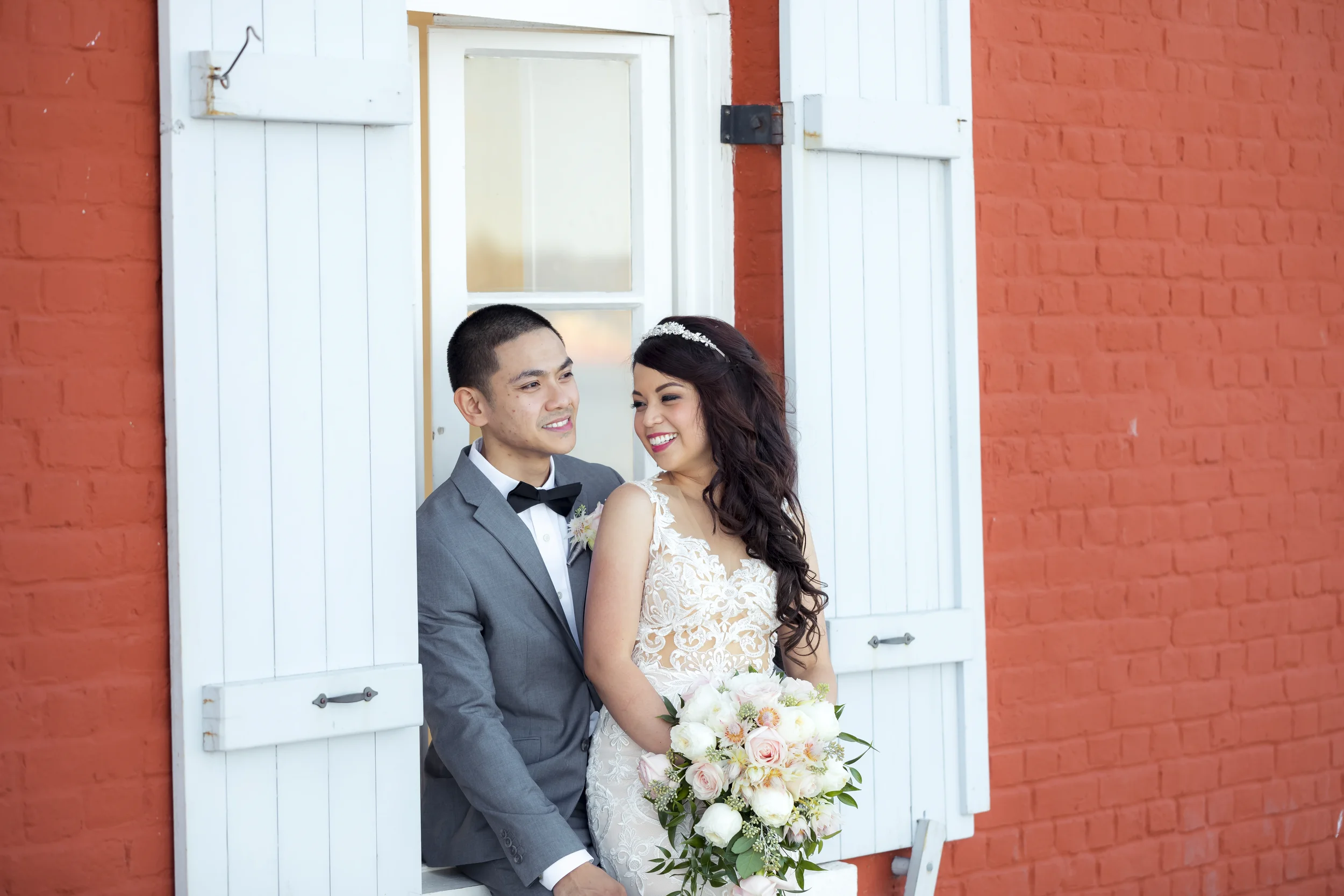 Smiling bride and groom posing together in an open white barn door frame against a red brick wall, bride in a lace wedding gown with long veil holding a soft pastel rose bouquet and groom in gray suit with black bow tie, captured in natural daylight,