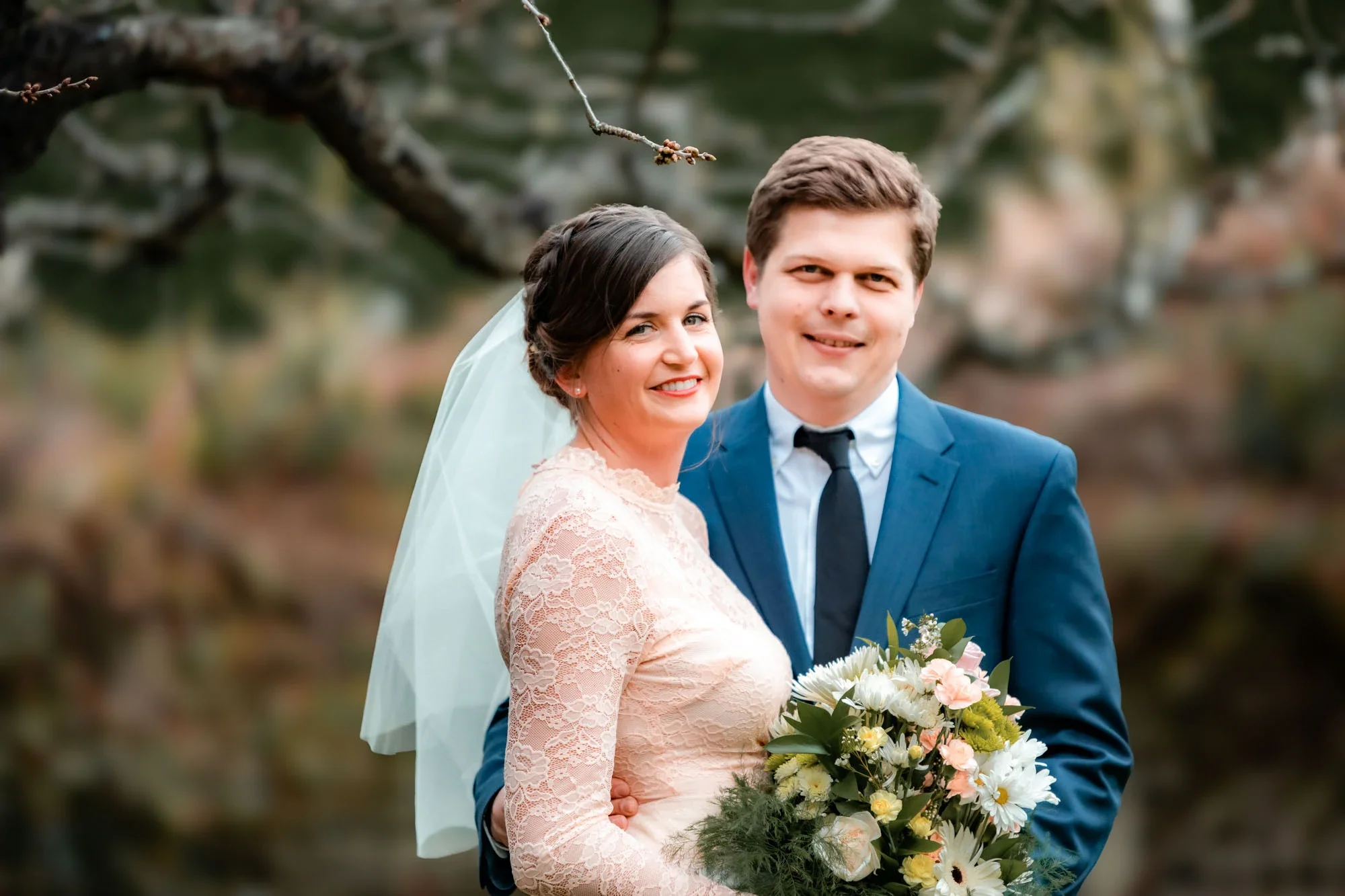 Joyful bride in a delicate blush pink lace wedding gown with veil and groom in navy suit smiling together during their outdoor wedding portrait session amidst spring blossoms in Abbotsford, British Columbia.