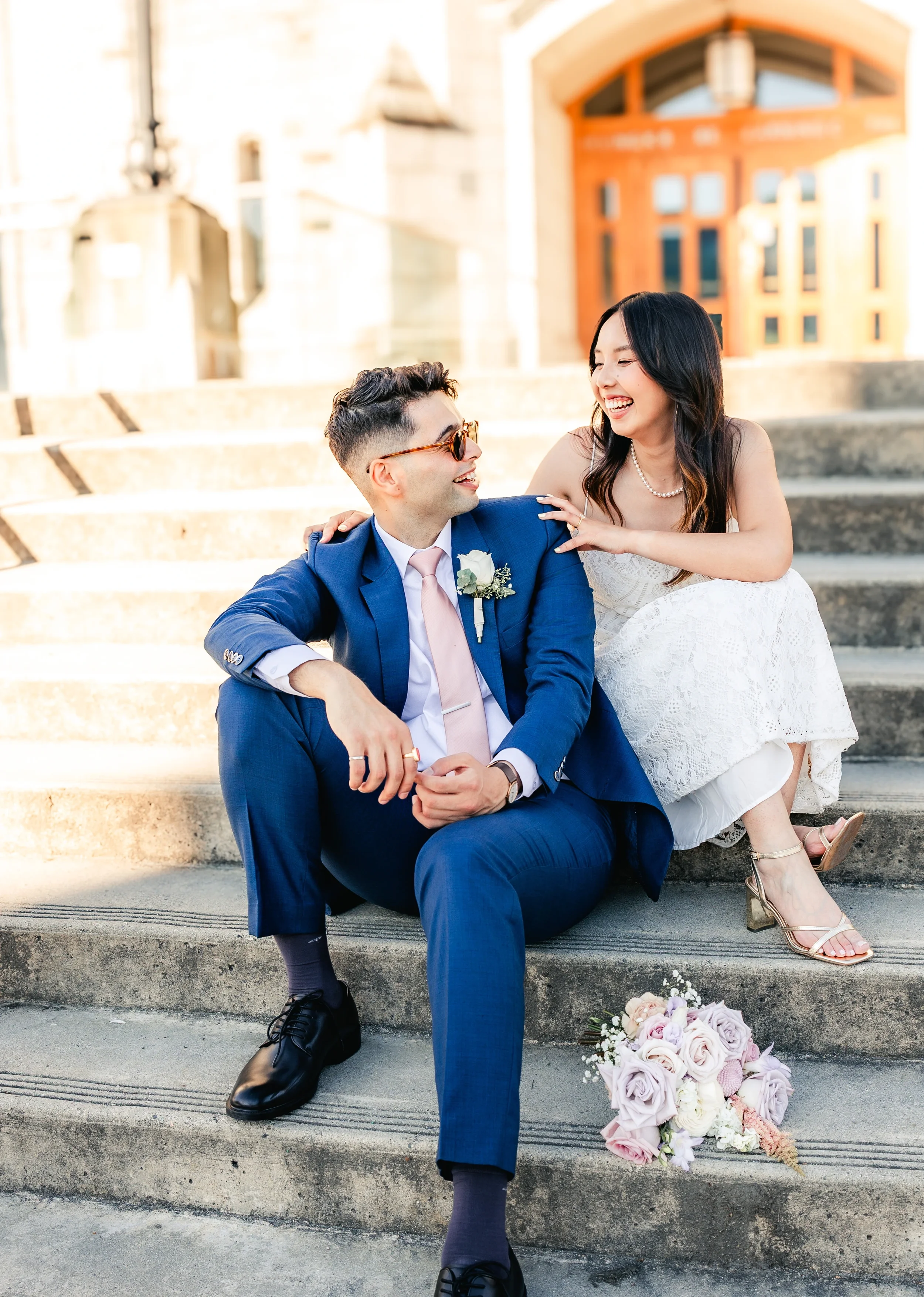 Happy bride and groom laughing together while sitting on steps of one of the libraries at the University of British Columbia, with groom in blue suit and pink tie, bride in elegant lace wedding dress holding a soft pink rose bouquet, captured by an a