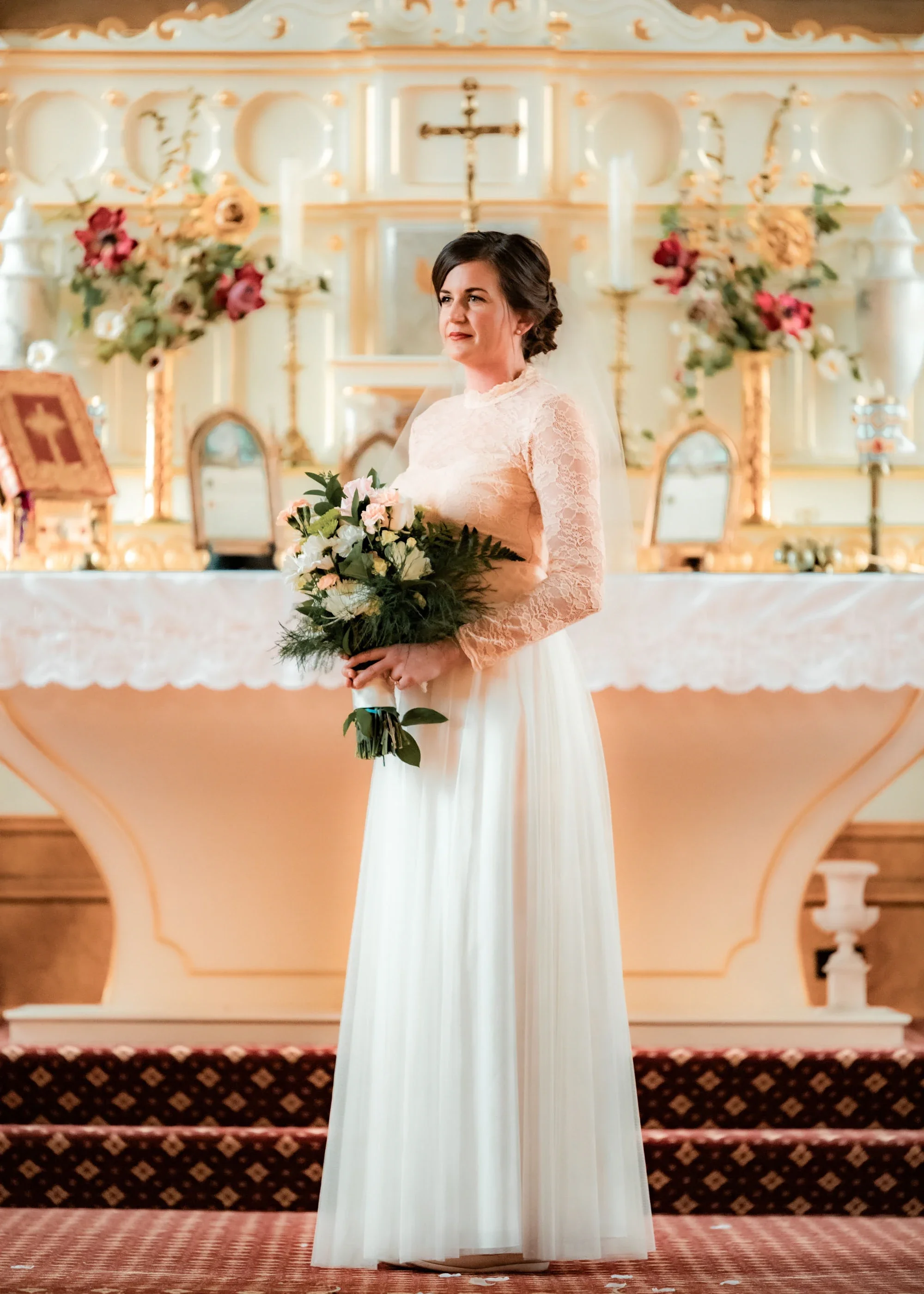 Elegant bride in a modest long-sleeve lace wedding dress holding a lush bouquet during an Orthodox Christian wedding ceremony in a beautifully adorned church with golden iconostasis, captured by an Abbotsford Fraser Valley wedding photographer.