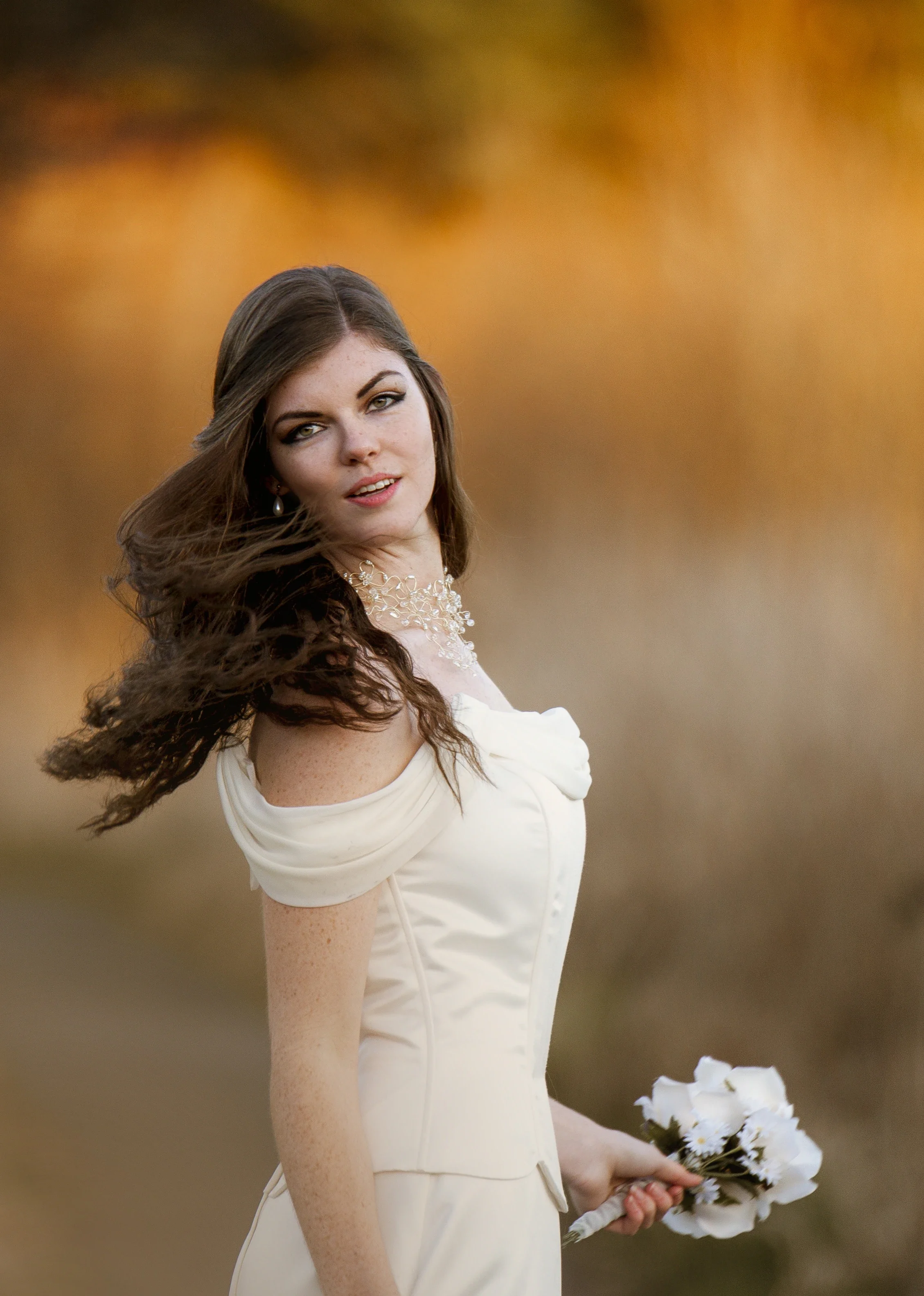 Radiant bride Megan with wind-swept hair looking back over her shoulder during golden hour, wearing an elegant off-the-shoulder wedding gown and holding a white daisy bouquet against warm blurred autumn tones, affordable wedding photography by Shakil