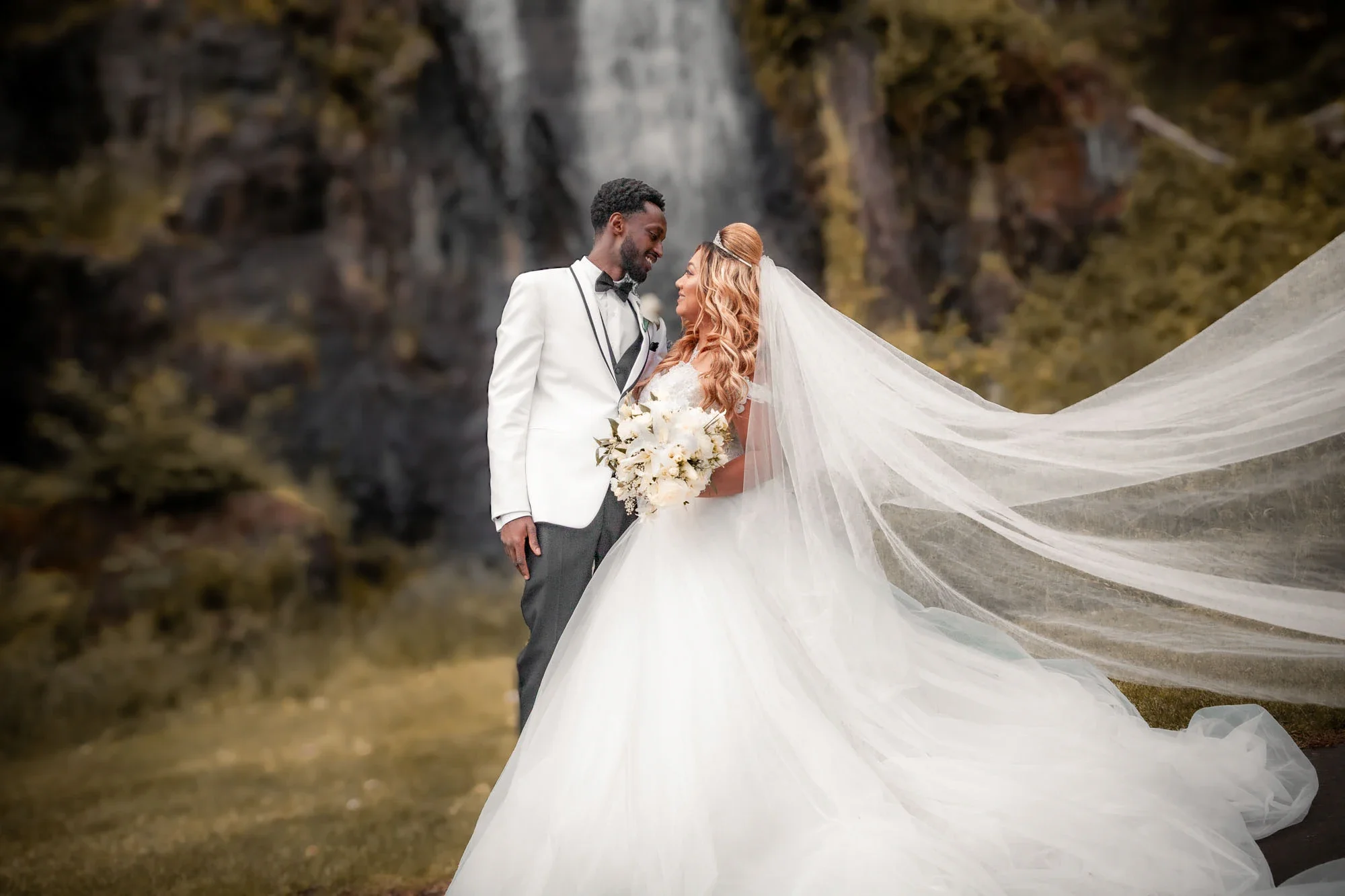 Tender moment as bride and groom gaze lovingly into each other's eyes with flowing veil catching the breeze, groom in white tuxedo jacket and bride in elegant ballgown holding a white orchid bouquet, framed by a misty waterfall and lush greenery, aff
