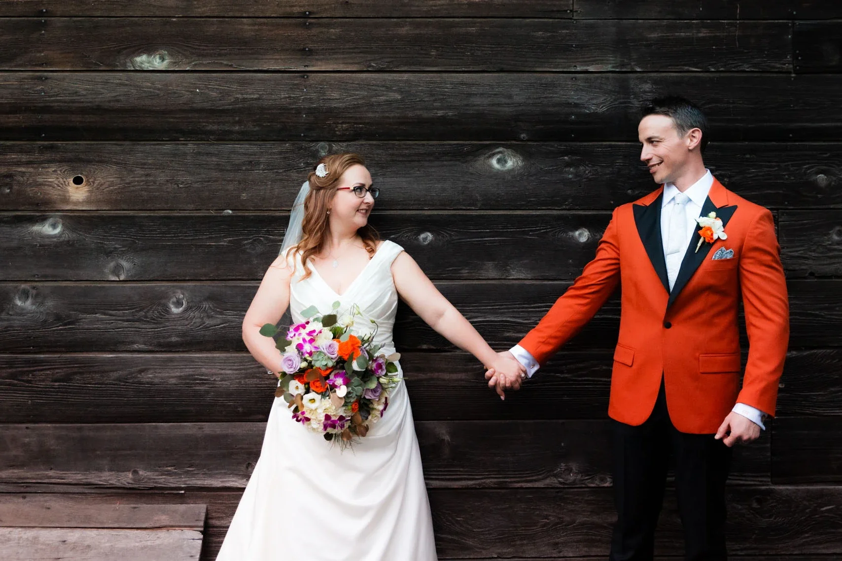 A bride and groom holding hands and smiling at each other in front of a wooden wall. The bride has red hair, glasses, a white dress, and is holding a colorful bouquet. The groom has short dark hair, is wearing a red jacket with black lapel, white shirt, and bow tie.