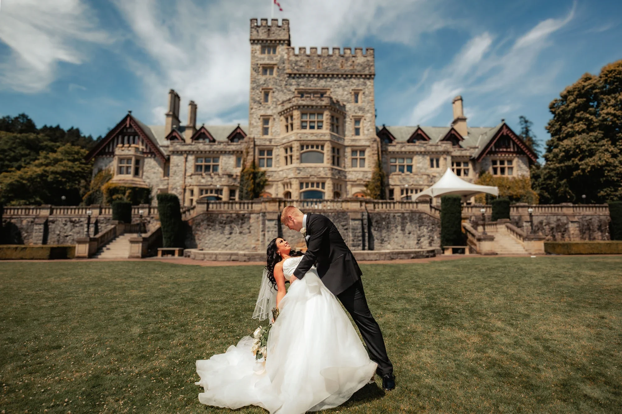 Playful groom dipping and kissing bride in front of majestic Hatley Castle with its iconic stone tower and Canadian flag, bride in flowing tulle wedding gown holding bouquet and groom in classic black suit, affordable wedding photography by Shakil Hu
