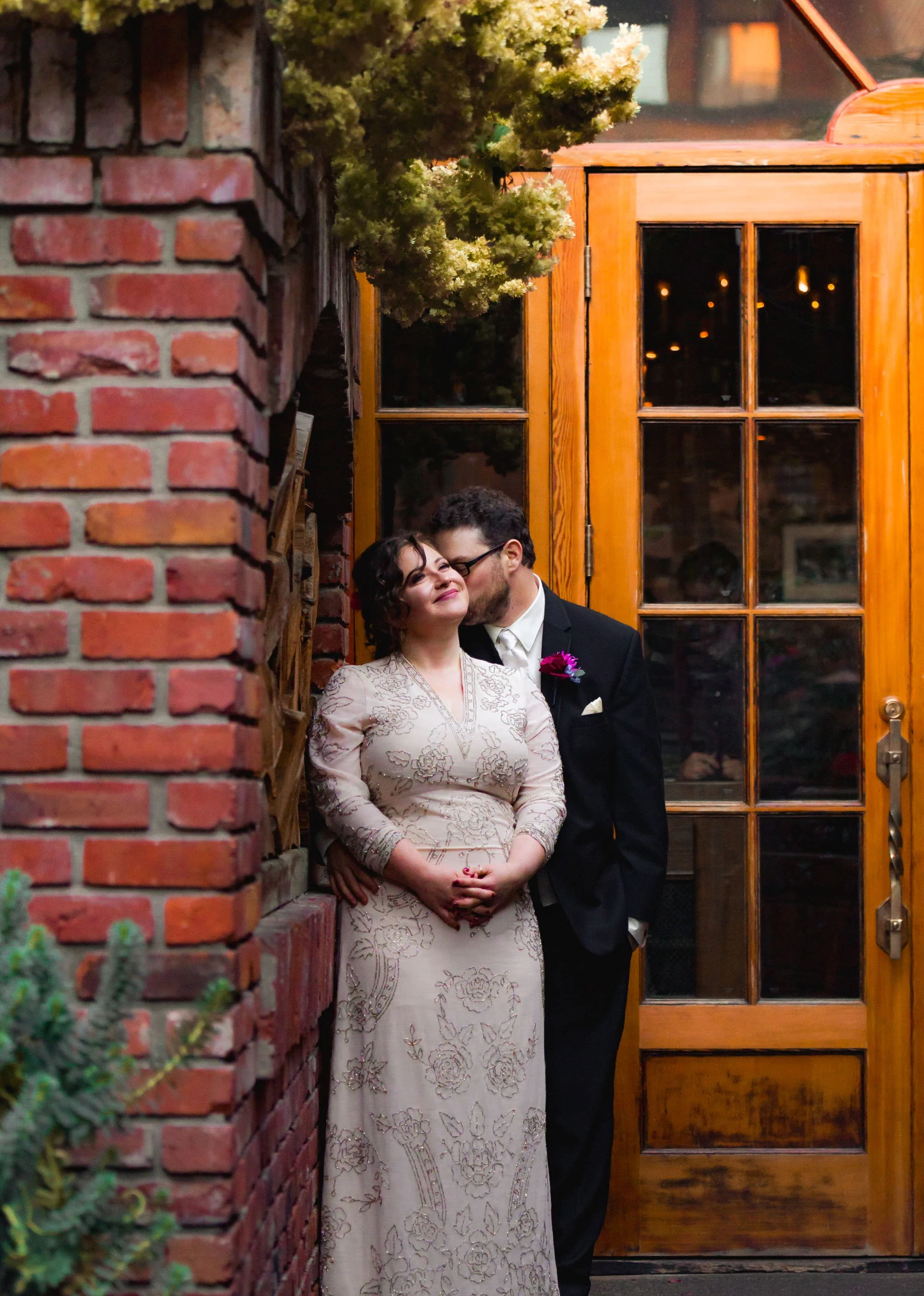 Tender moment as groom kisses bride on the forehead against a rustic red brick wall beside a charming wooden door with warm lighting, bride in elegant long-sleeve embroidered gown and groom in classic black suit, beautifully captured by Shakil Hussai