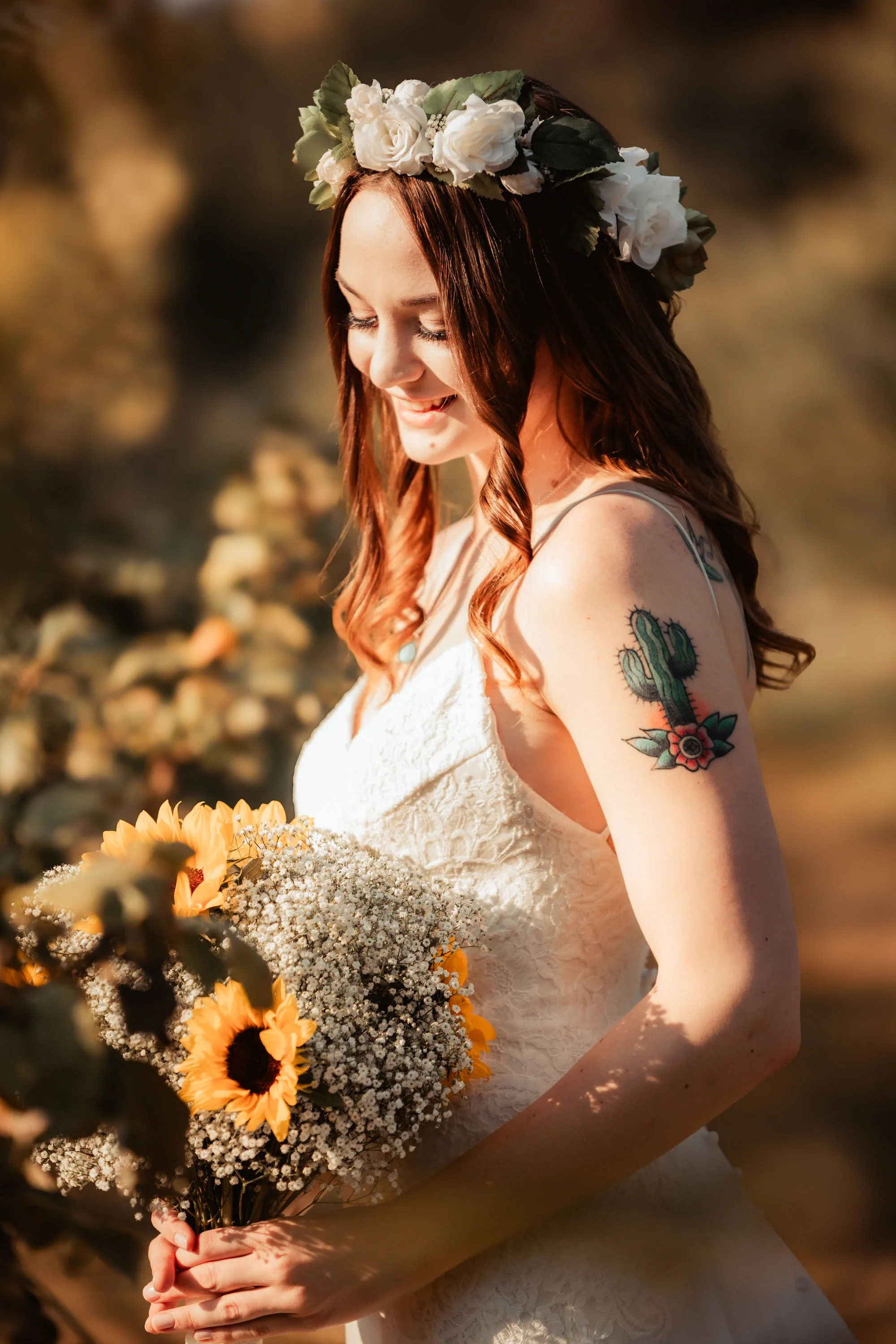 Boho bride with floral crown and cactus tattoo admiring her vibrant sunflower and baby's breath bouquet during golden hour in a natural outdoor setting, wearing a textured lace wedding gown, affordable wedding photography by Shakil Hussain Photograph