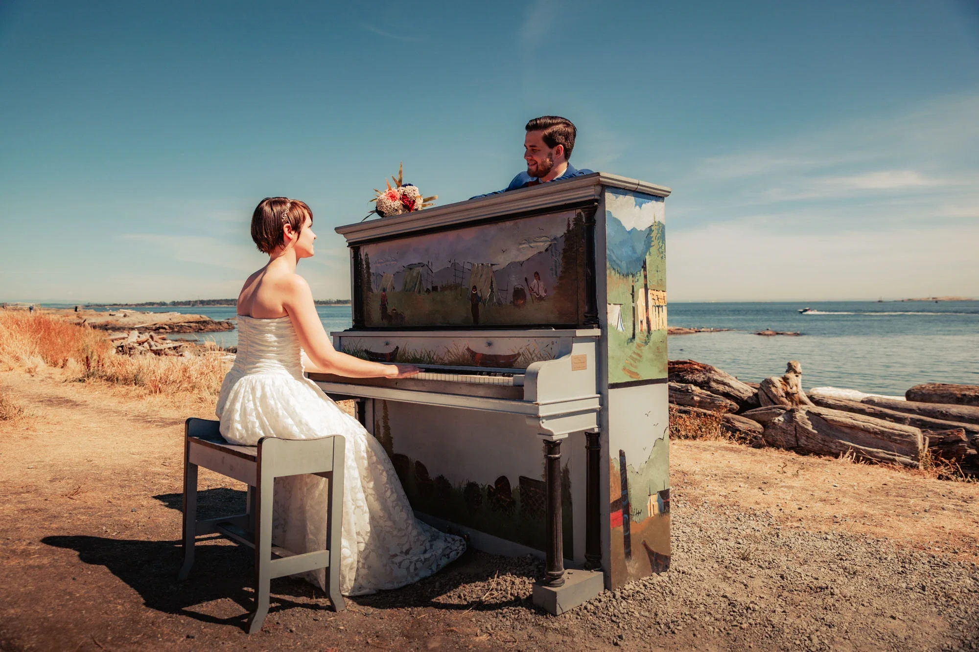 Creative wedding portrait of bride in strapless white gown playing a painted upright piano on the beach with bouquet resting on top, groom in blue suit smiling beside her against a serene ocean backdrop under clear skies, affordable wedding photograp
