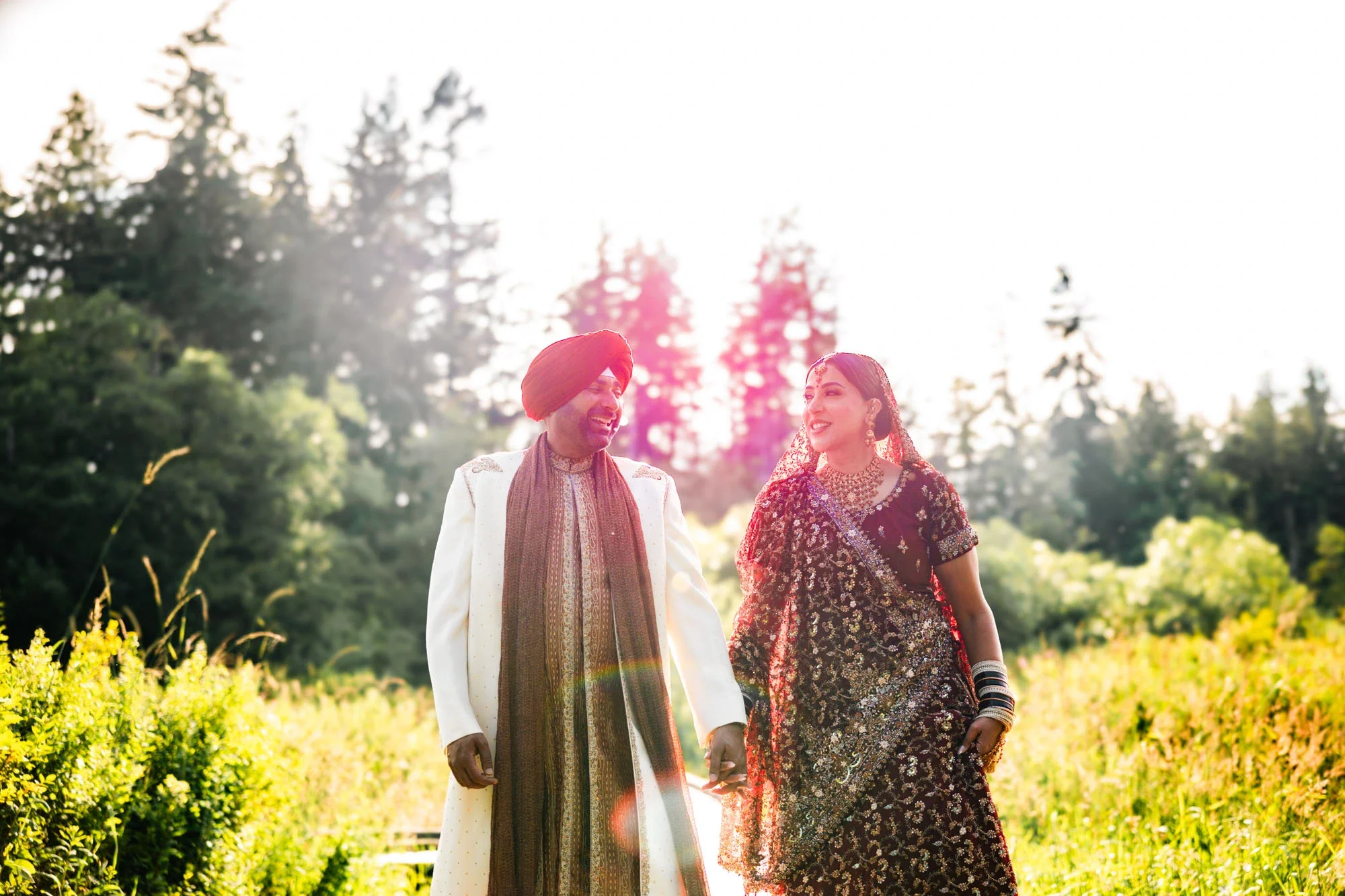 Bride and groom in traditional Indian attire walking hand-in-hand through a sunlit field with golden hour lens flare and lush greenery, groom in cream sherwani with red turban and bride in ornate maroon embroidered lehenga, affordable wedding photogr