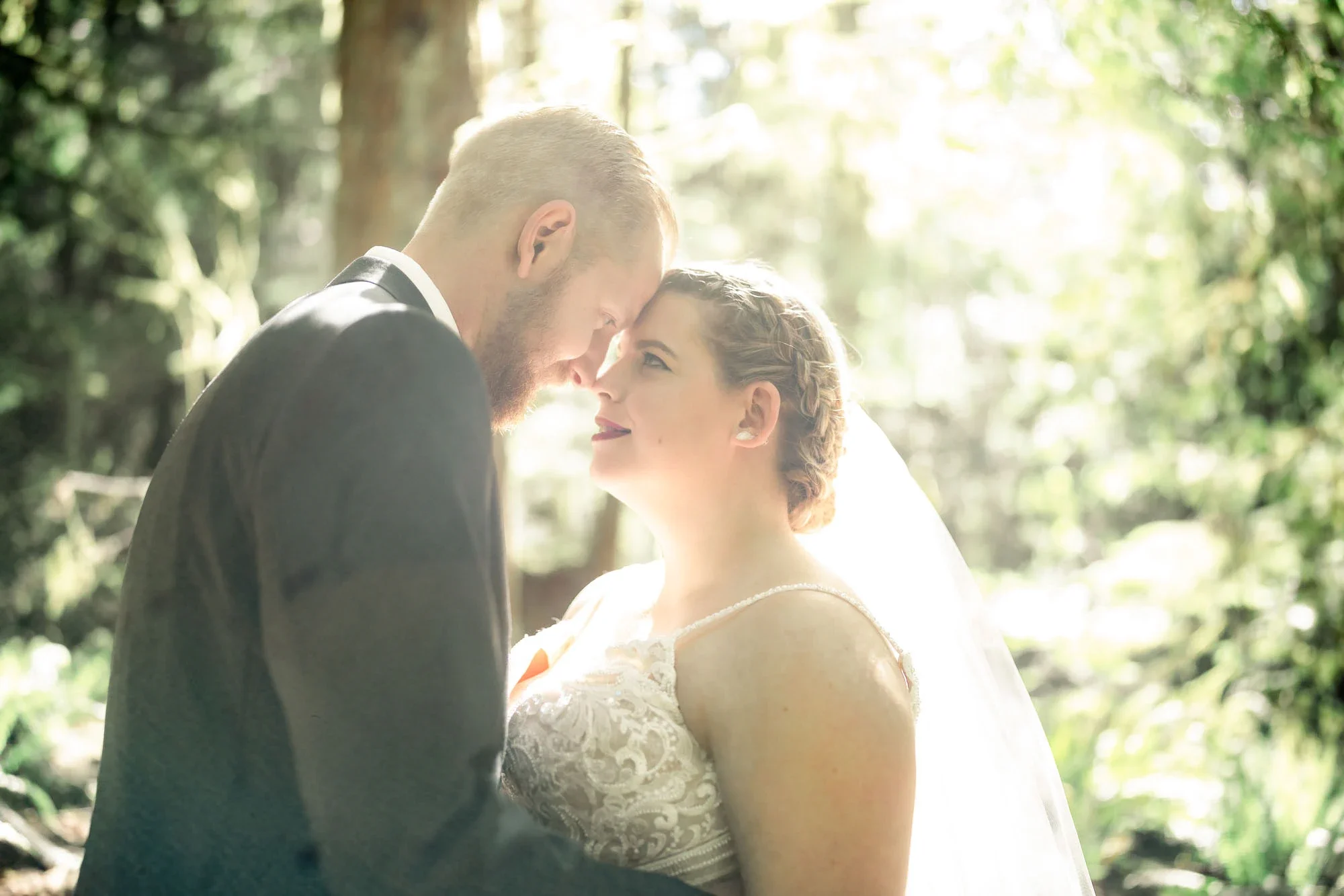 Intimate moment as bride and groom touch foreheads in a sunlit forest, bride in a detailed lace wedding gown with veil and groom in gray suit sharing a tender connection with soft bokeh greenery in the background, affordable wedding photography by Sh
