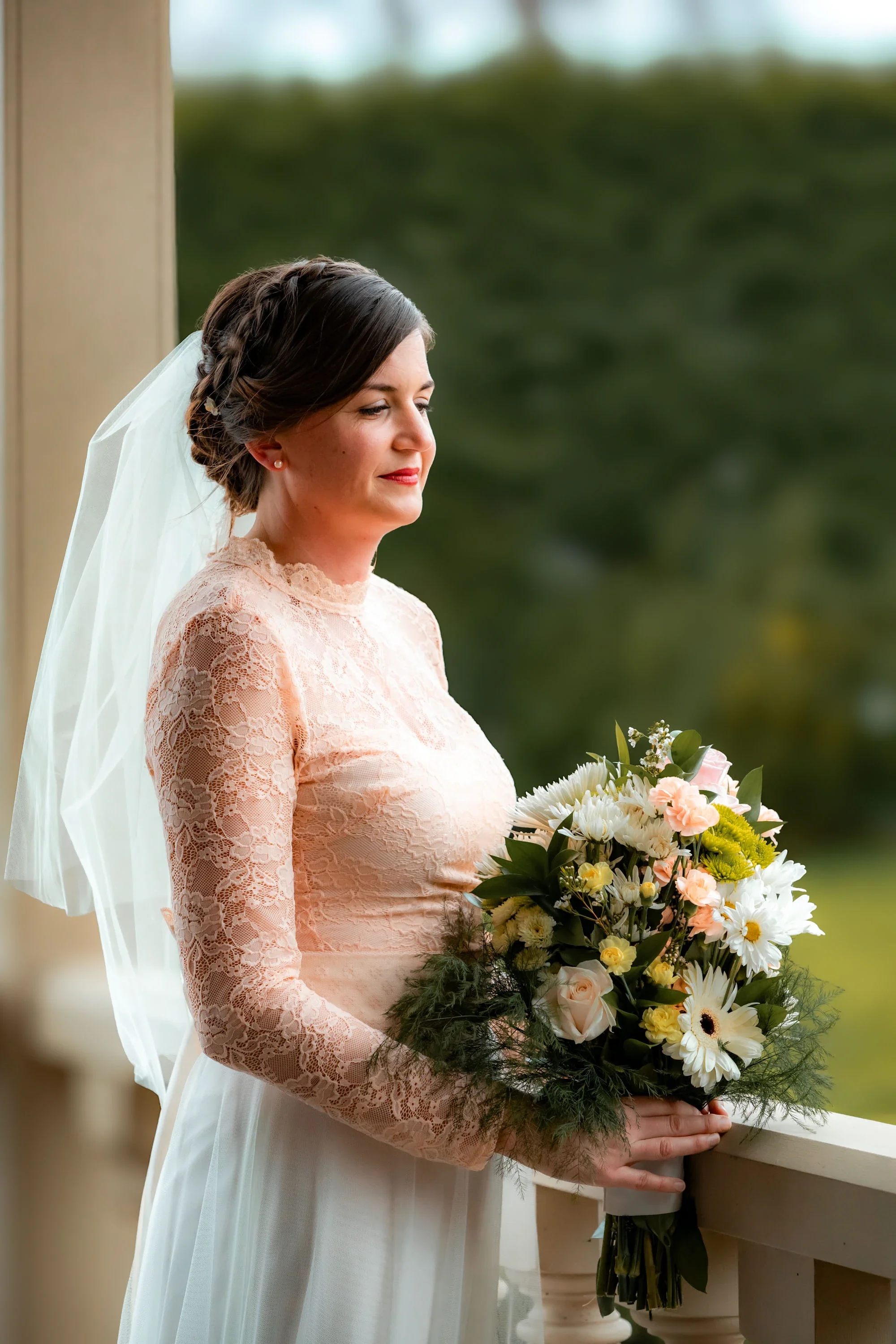 Elegant bride in a long-sleeved blush lace wedding dress holding a lush bouquet of white, peach, and green flowers, smiling softly outdoors with a blurred natural background in Abbotsford BC. Captured by Shakil Hussain, Abbotsford BC wedding photogra