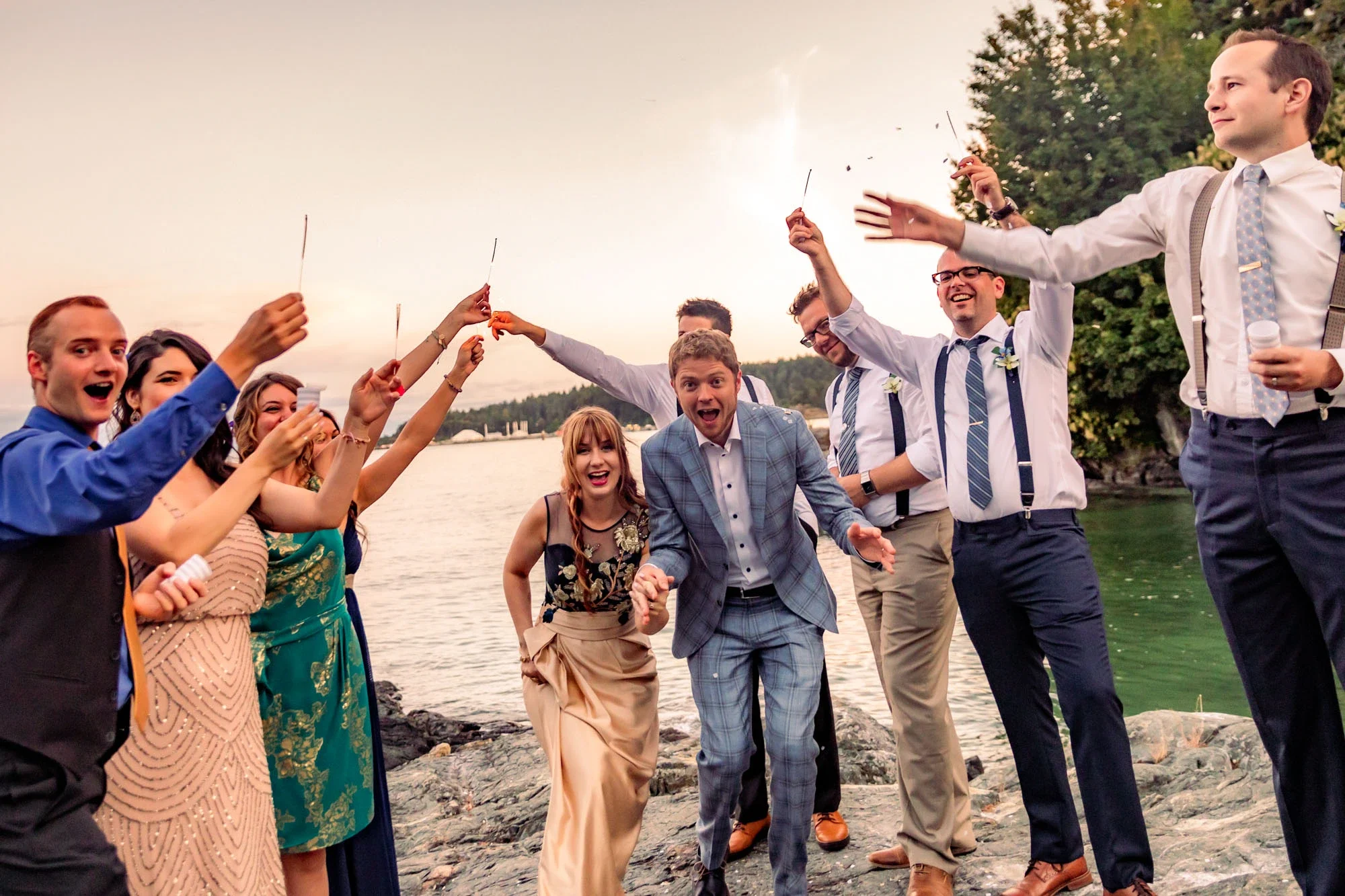 Excited wedding party celebrating with sparklers during a joyful send-off at sunset by the lake in Abbotsford BC, with the bride and groom in the center surrounded by cheering friends. Captured by Shakil Hussain, Abbotsford BC wedding photographer.