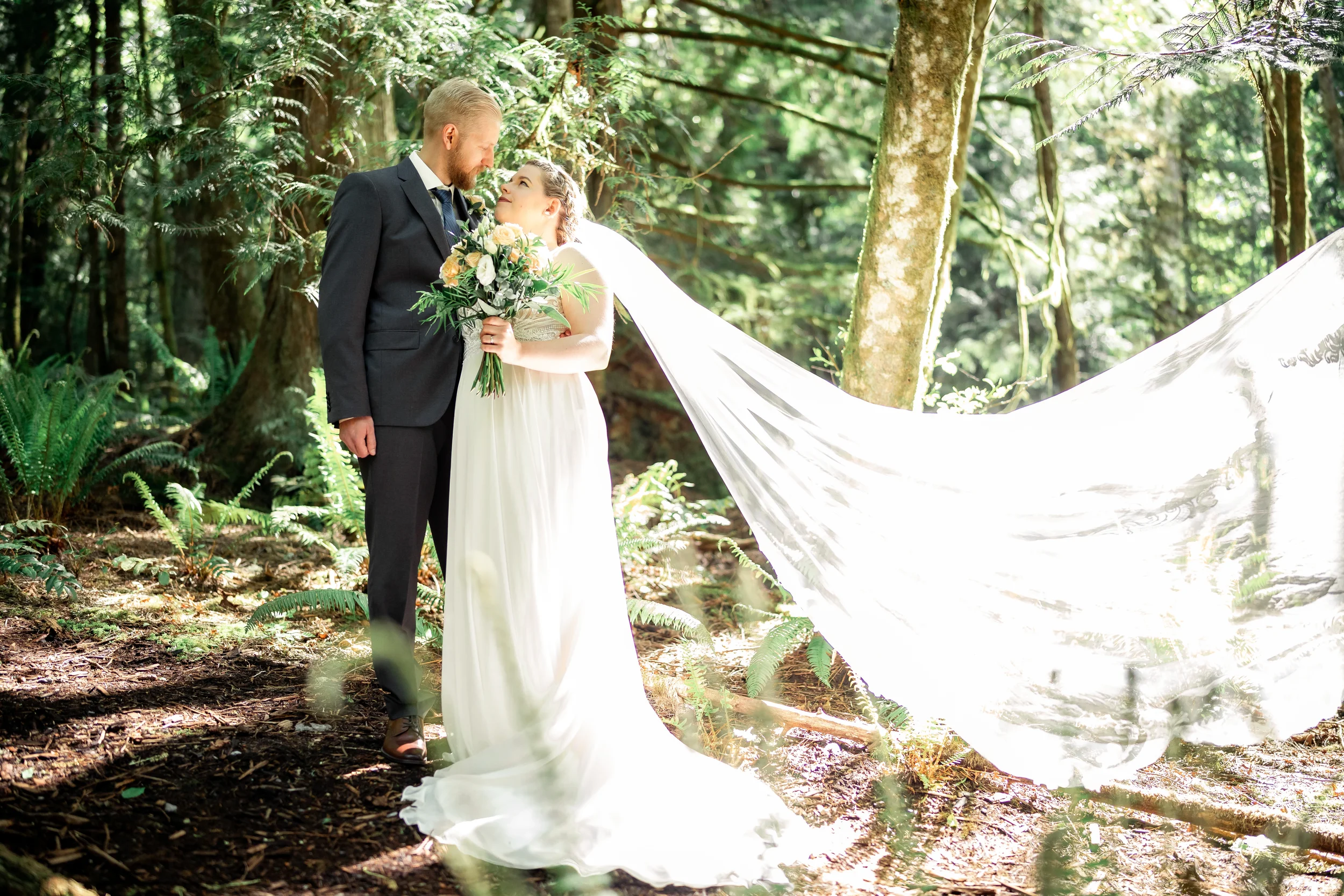 Romantic bride and groom sharing a quiet moment in a lush Pacific Northwest forest, with dramatic long flowing veil catching the sunlight and bride holding a natural peach rose bouquet while groom in dark suit stands close behind her, affordable wedd