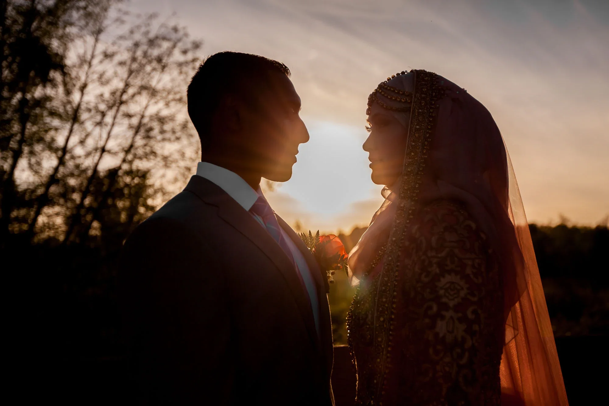 Dramatic silhouette of bride and groom gazing at each other during golden sunset, bride in traditional embroidered lehenga with veil and groom in suit with red rose boutonniere against a glowing sky and blurred trees, affordable wedding photography b