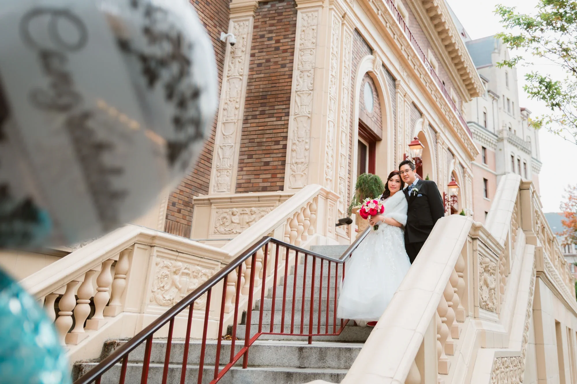 Happy bride and groom standing arm-in-arm on an impressive ornate stone staircase with intricate carvings and brick details at a historic venue, bride in sparkling ballgown with fur stole holding vibrant pink bouquet and groom in classic black tuxedo