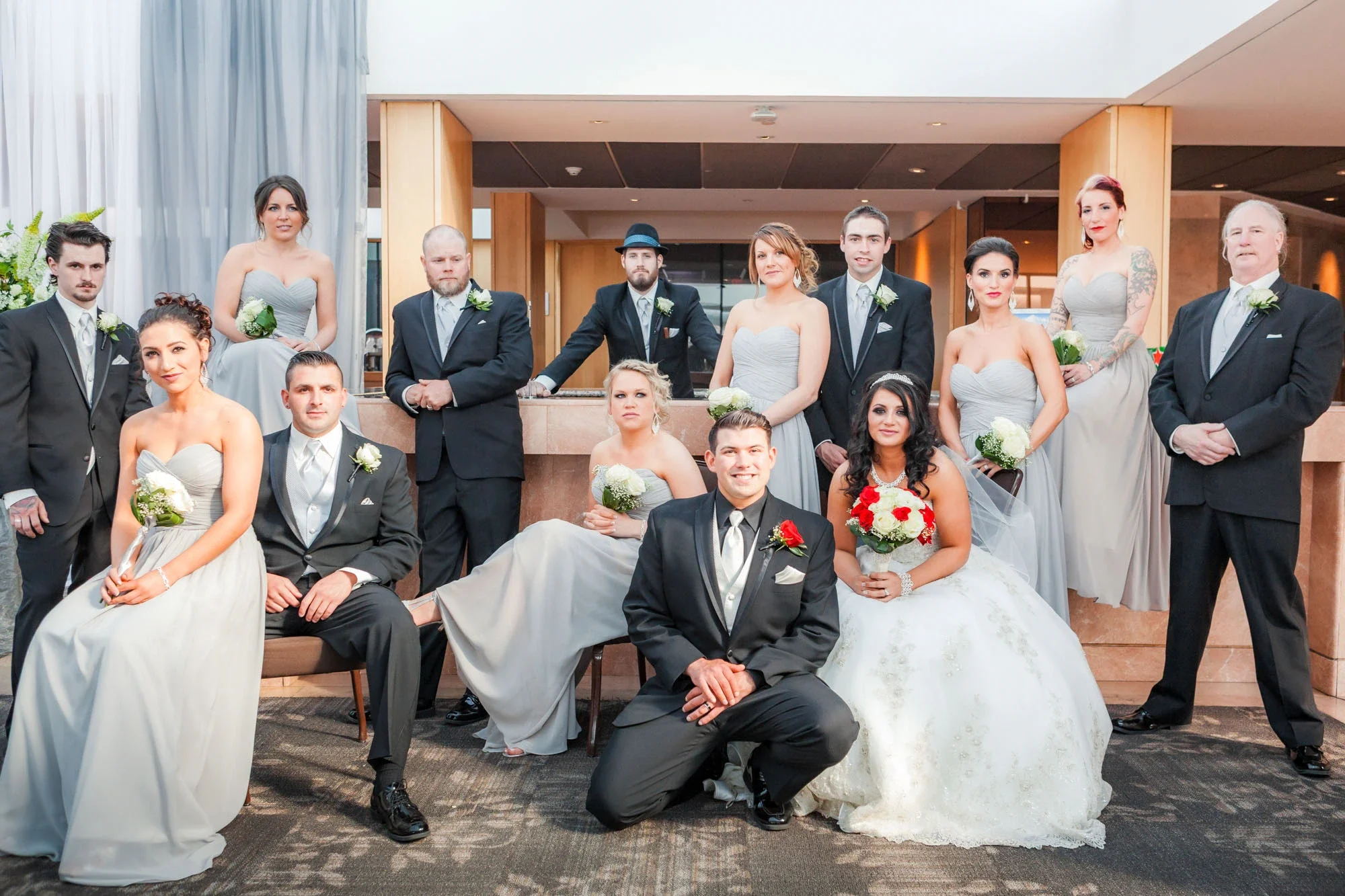 Elegant full wedding party group portrait featuring the bride in a classic white gown and groom in black tuxedo, surrounded by bridesmaids in light gray dresses and groomsmen in black suits. Captured indoors by Shakil Hussain, Abbotsford BC wedding p