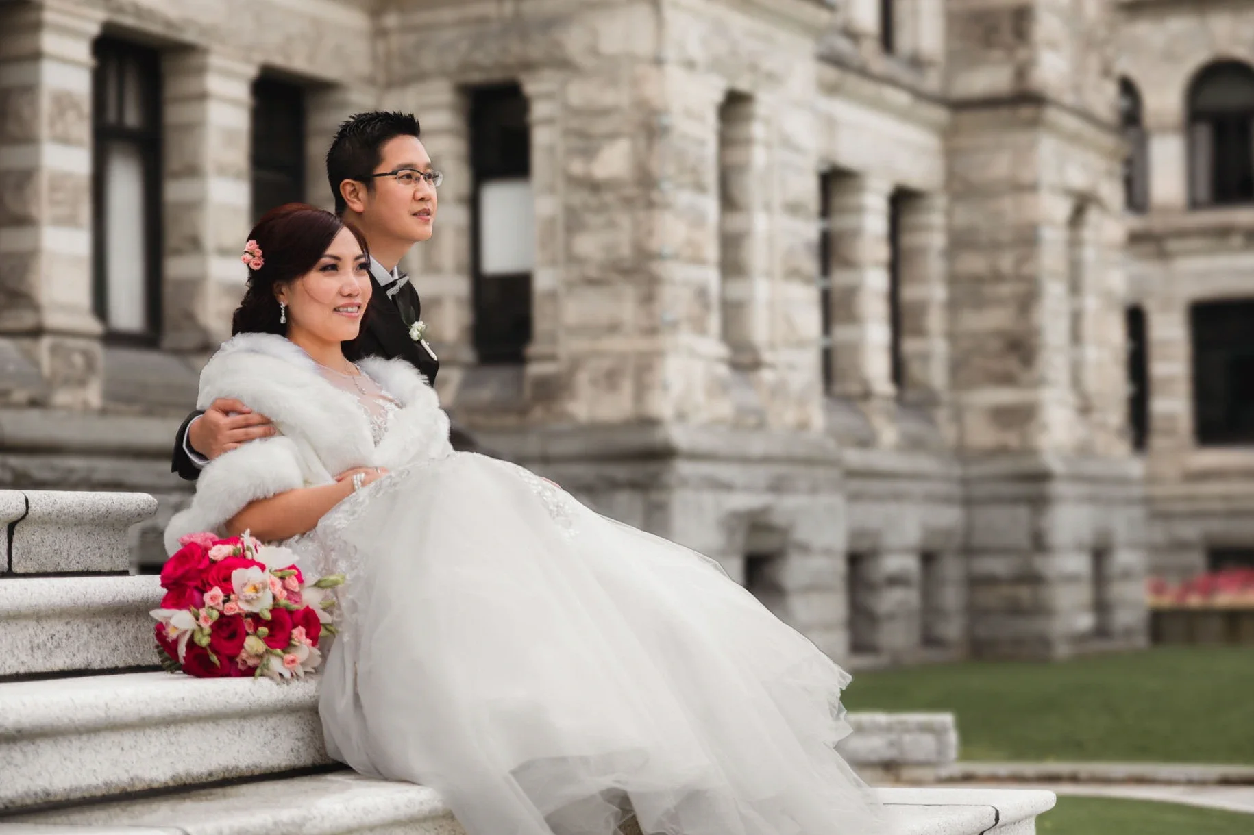 Bride and groom sitting on steps outside a stone building, the bride in a white wedding gown with a fur stole and the groom in a black tuxedo. The bride has a bouquet of red and pink flowers resting on the steps beside her.