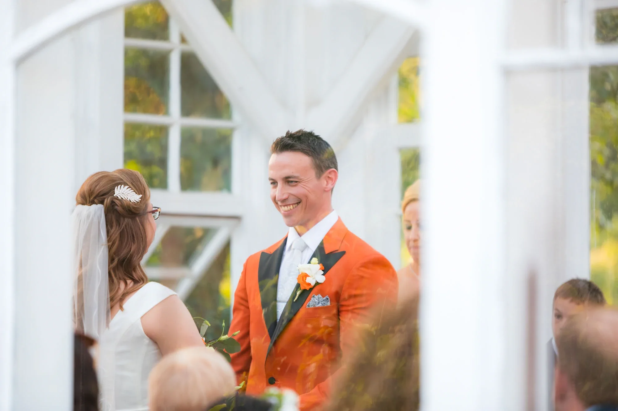 Joyful groom in vibrant orange suit with black lapels and orange rose boutonniere smiling during the outdoor wedding ceremony in a white gazebo, with bride seen from behind holding bouquet, affordable wedding photography by Shakil Hussain Photography