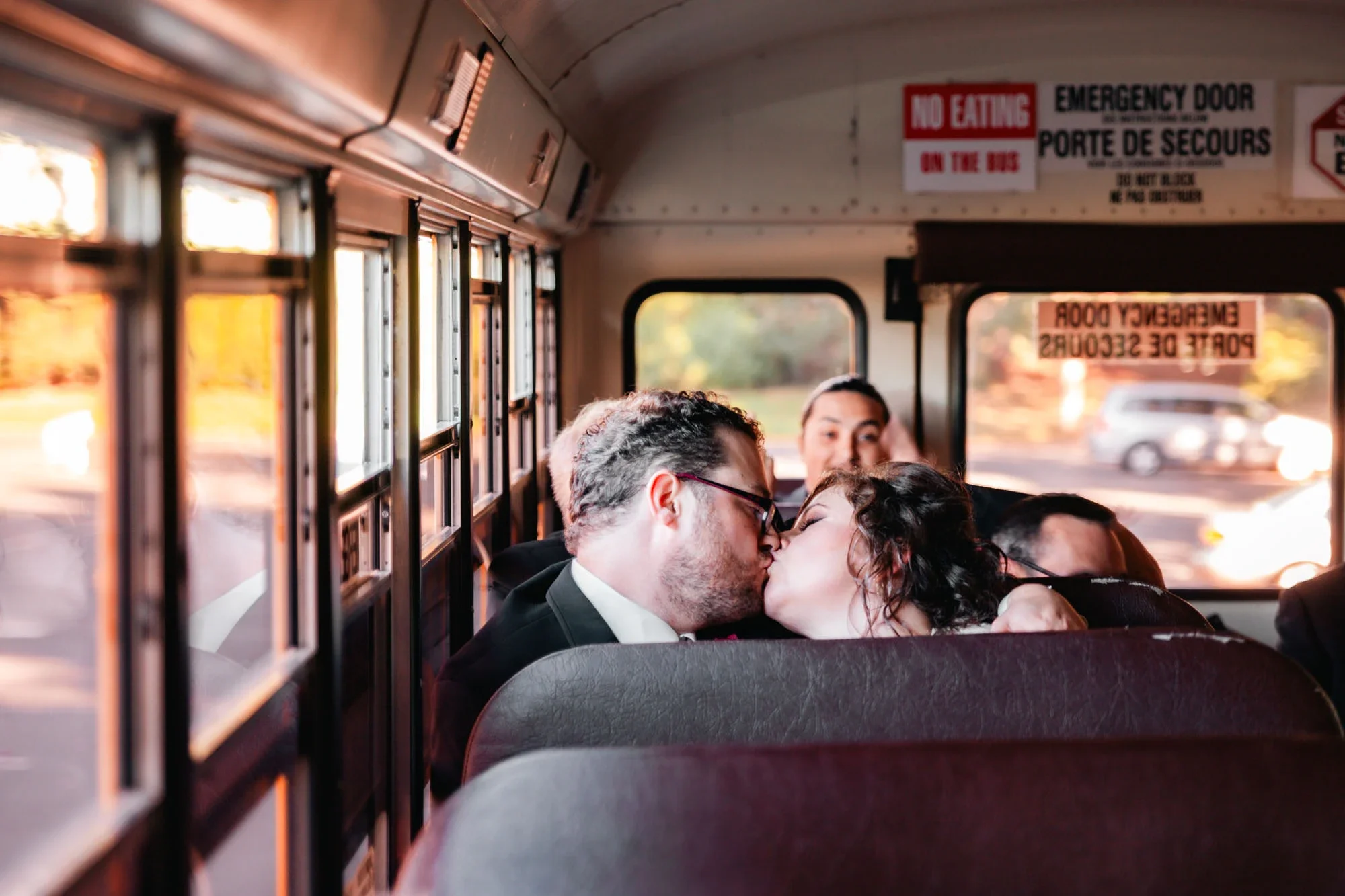 A couple kissing on a school bus with other passengers and a window view of roads and cars.
