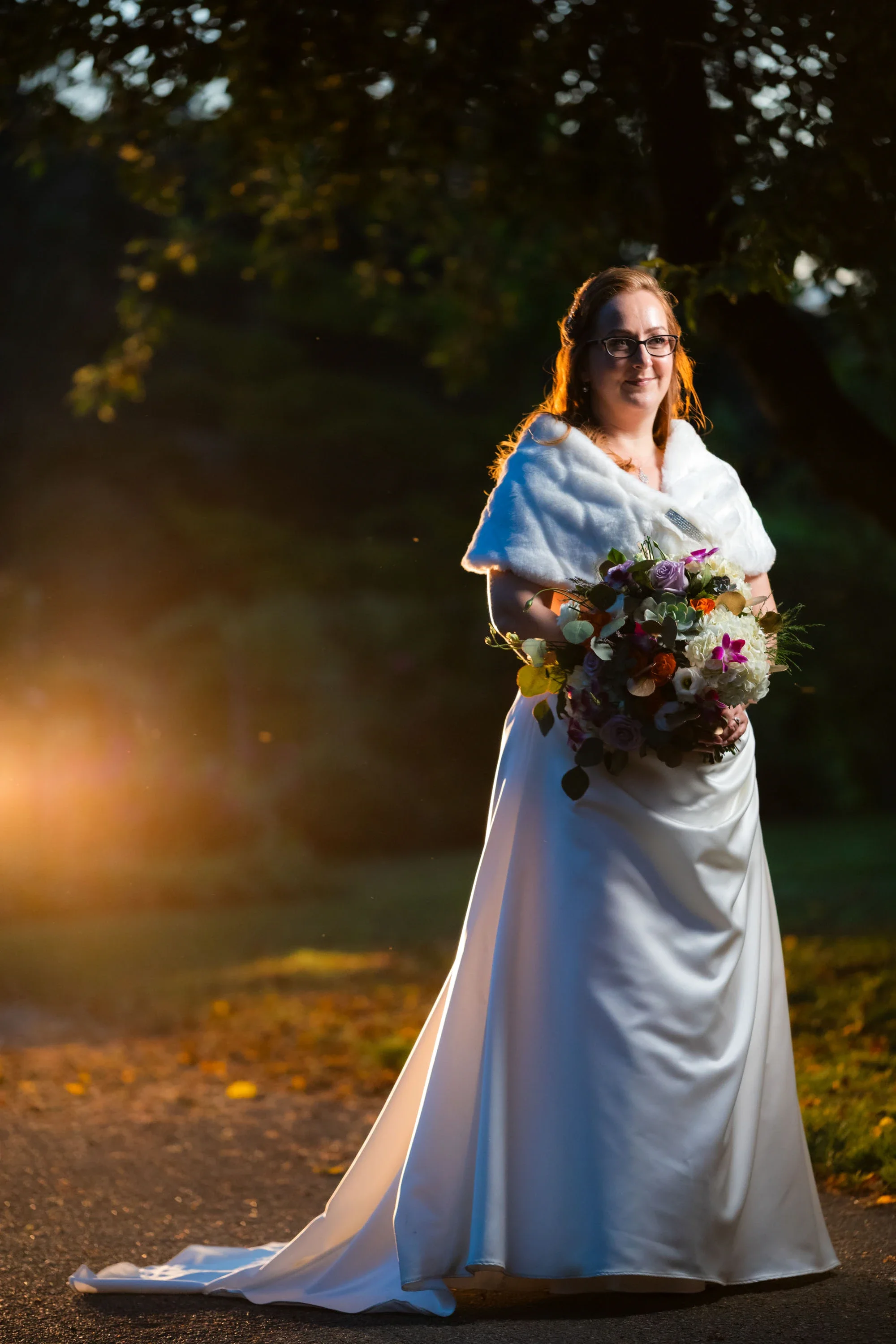 Elegant bride in a classic satin wedding gown with white fur stole holding a vibrant mixed bouquet with succulents, roses, and orchids, smiling softly during golden hour on a tree-lined path, affordable wedding photography by Shakil Hussain Photograp