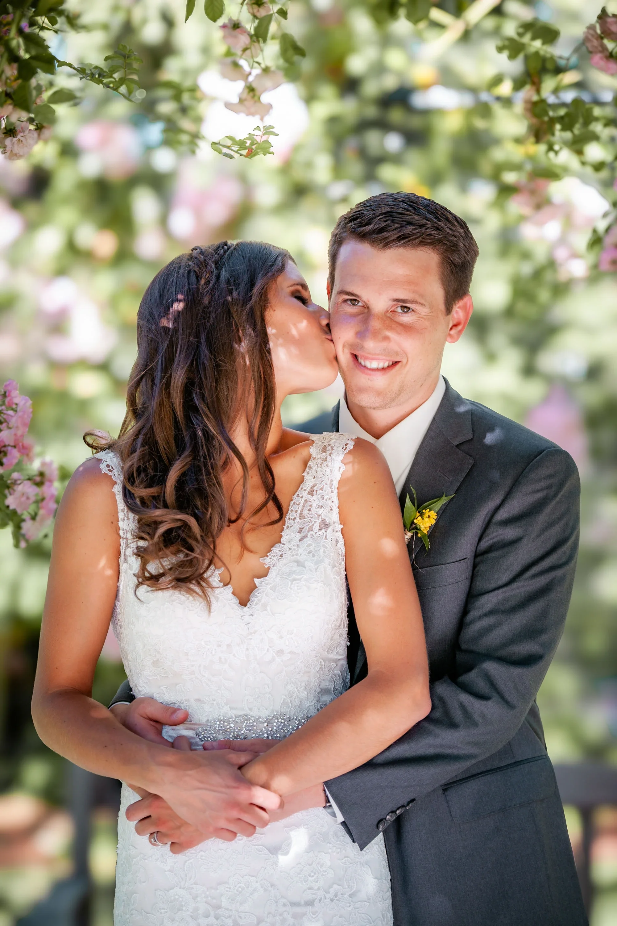 Sweet moment as bride kisses groom on the cheek while embraced in front of soft pink spring blossoms, bride in elegant lace wedding gown with beaded belt and groom in charcoal suit with yellow boutonniere, affordable wedding photography by Shakil Hus