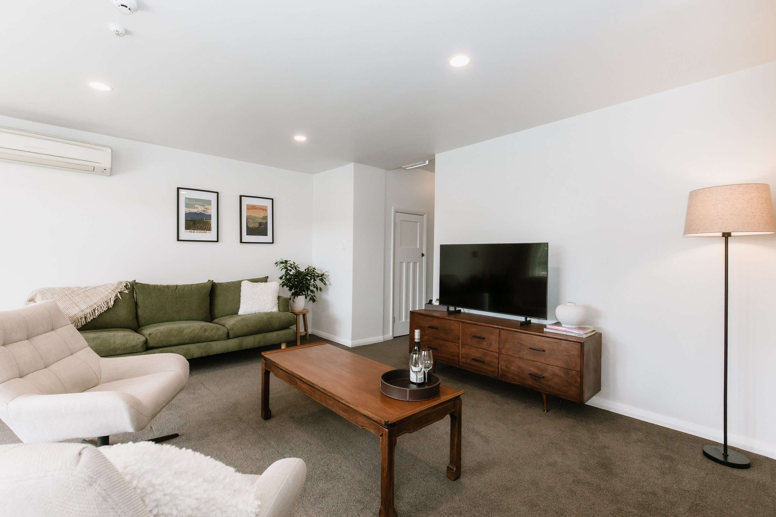 Living room with a beige armchair, a green sofa, a wooden coffee table with bottles and glasses, a wooden TV stand with a large flat-screen TV, a white vase, a tall floor lamp, framed artwork, and potted plants against white walls.