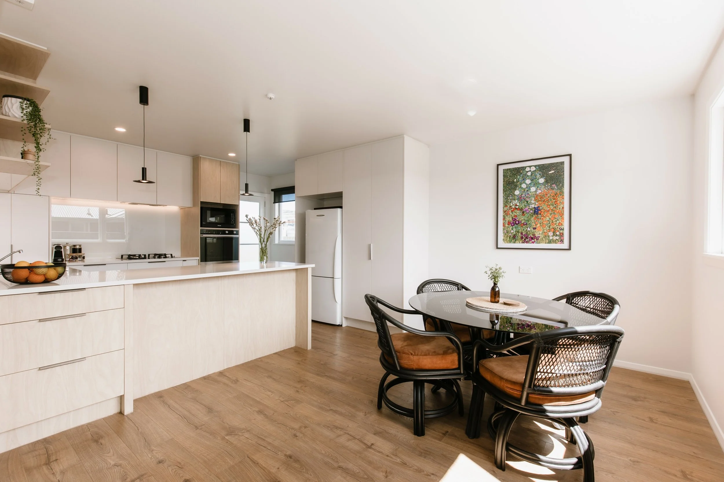 Modern kitchen and dining area with light wood cabinetry, black pendant lights, a white refrigerator, a round dining table with six black chairs, and a colorful floral art piece on the wall.