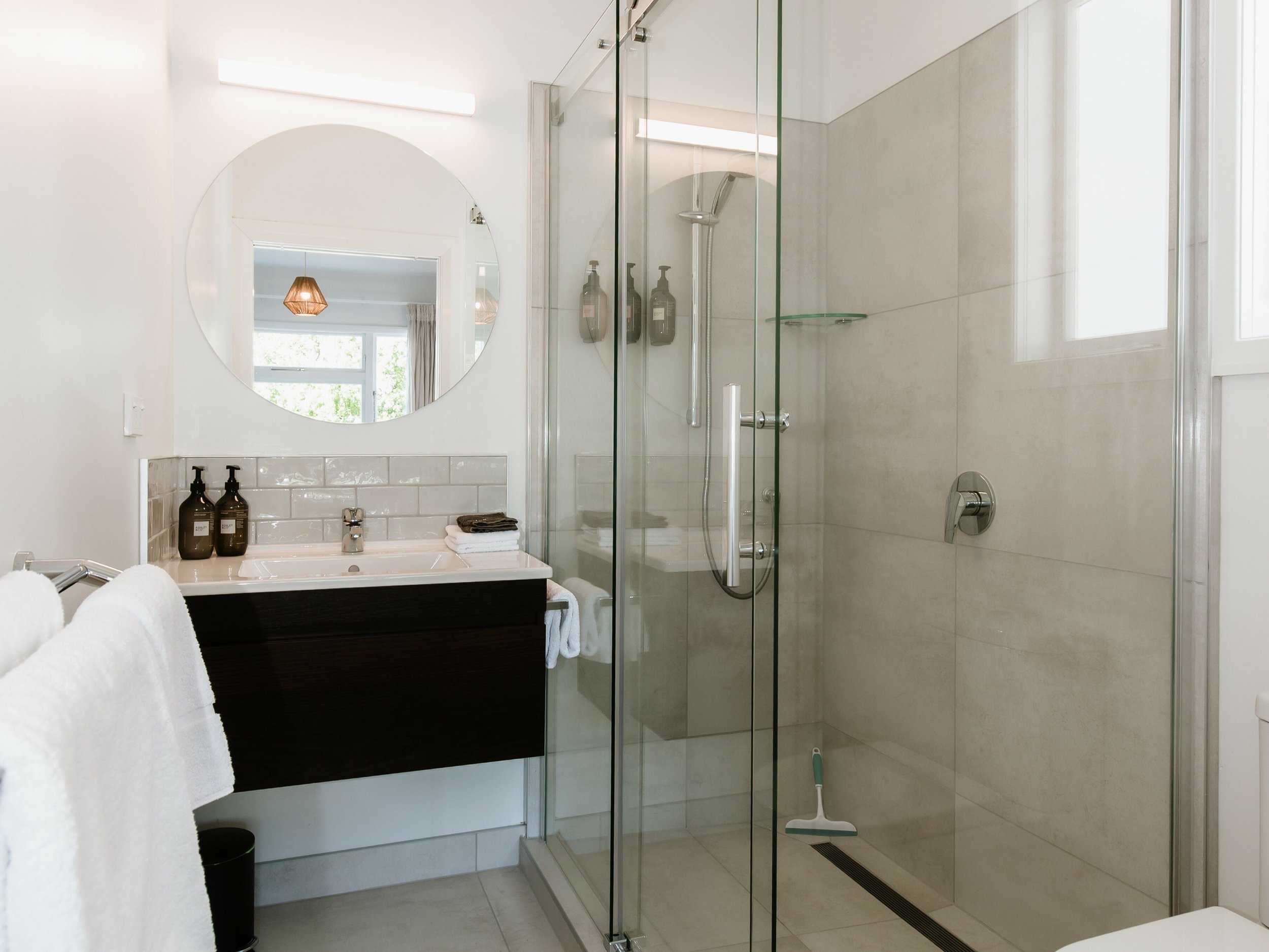 Modern bathroom with a glass-enclosed shower, a white sink with two soap dispensers, a round mirror, towels, and a window providing natural light.